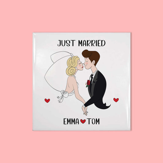 Vierkant keramisch tegeltje met bruid en bruidegom in cartoonstijl, met de tekst 'Just Married' en namen Emma & Tom, gefotografeerd recht van voren op roze achtergrond.