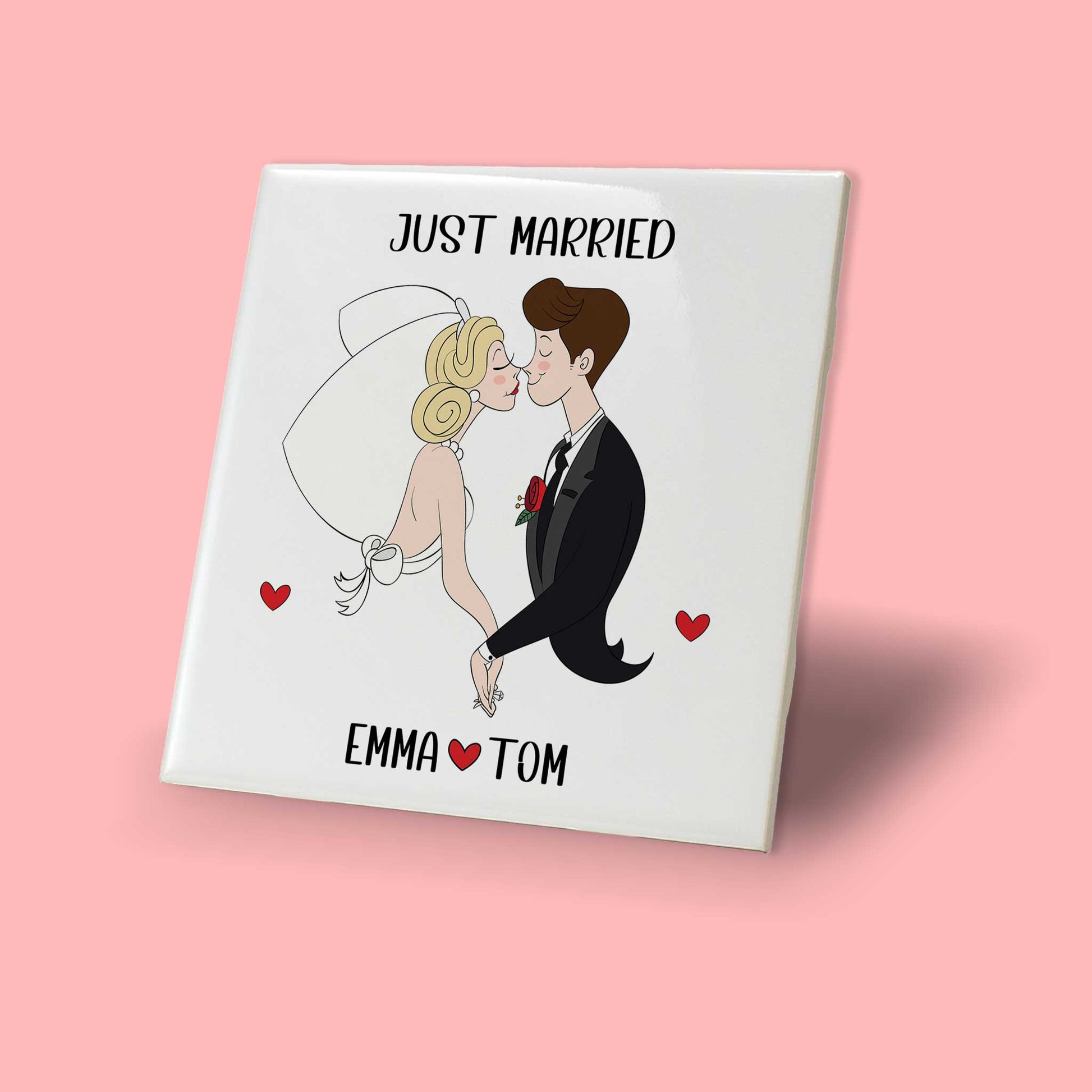 Wit keramisch trouwtegeltje met cartoonillustratie van een pasgetrouwd stel, tekst 'Just Married' erboven en namen Emma & Tom met hartje eronder, weergegeven tegen een roze achtergrond.