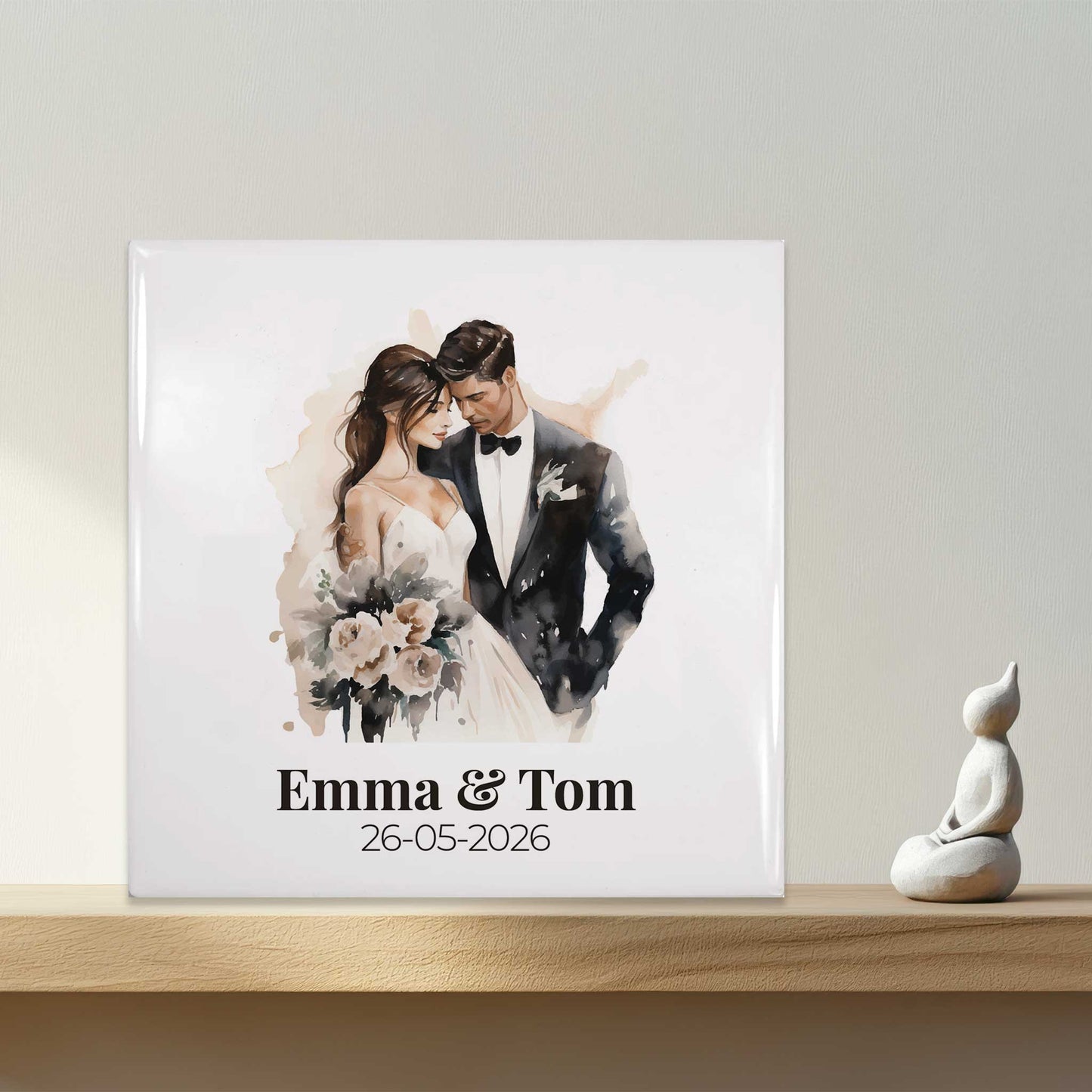 Vooraanzicht van een wit keramisch trouwtegeltje met illustratie van een bruidspaar, namen en trouwdatum, rechtopstaand op een houten plank met zacht licht van rechts.