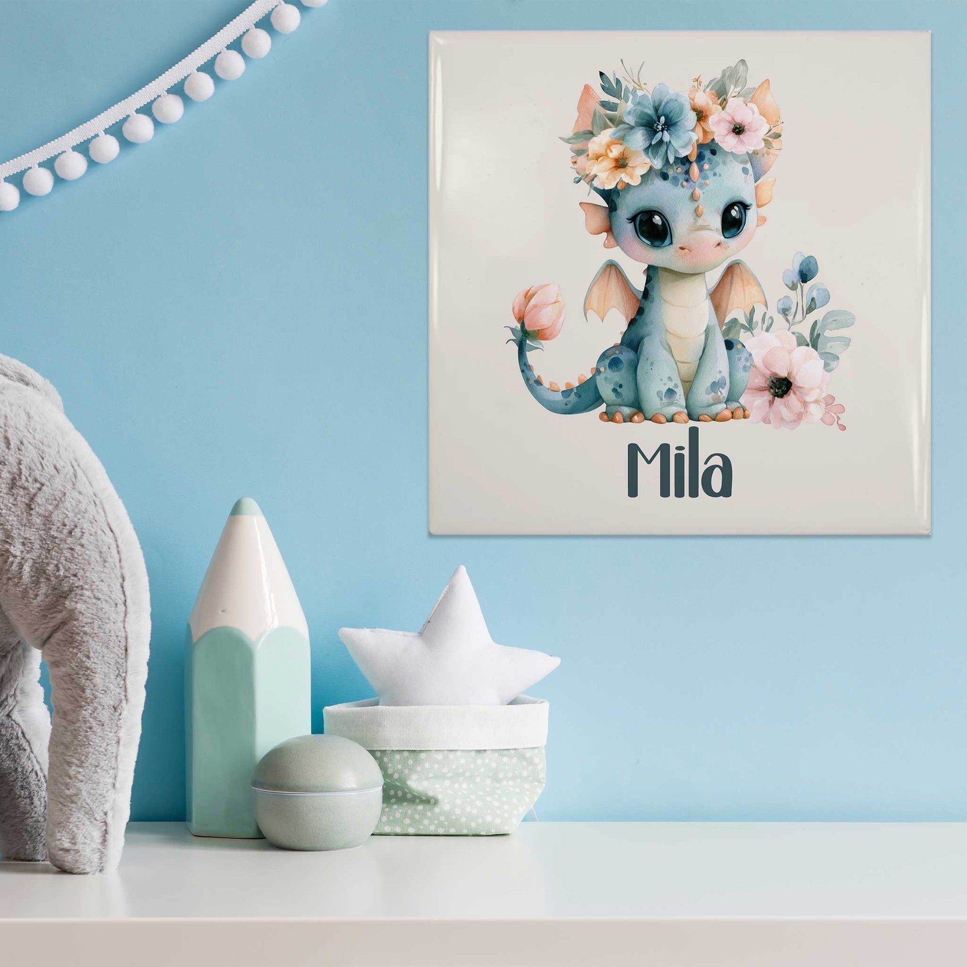 Mat keramisch tegeltje met een blauw draakje met bloemenkroon en de naam Mila aan een lichtblauwe muur boven een witte kast met babydecoratie.