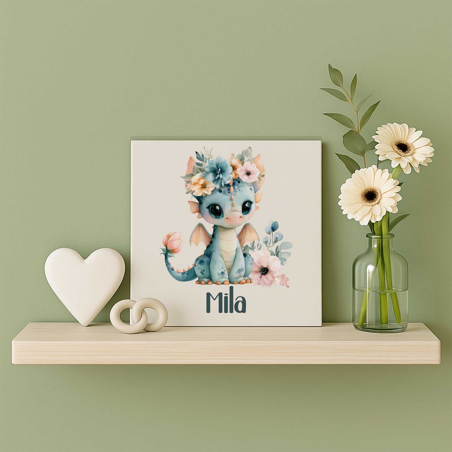 Mat keramisch tegeltje met een blauw draakje met bloemenkroon en de naam Mila op een houten wandplank naast een wit hart en een glazen vaasje met witte bloemen tegen een zachtgroene muur.