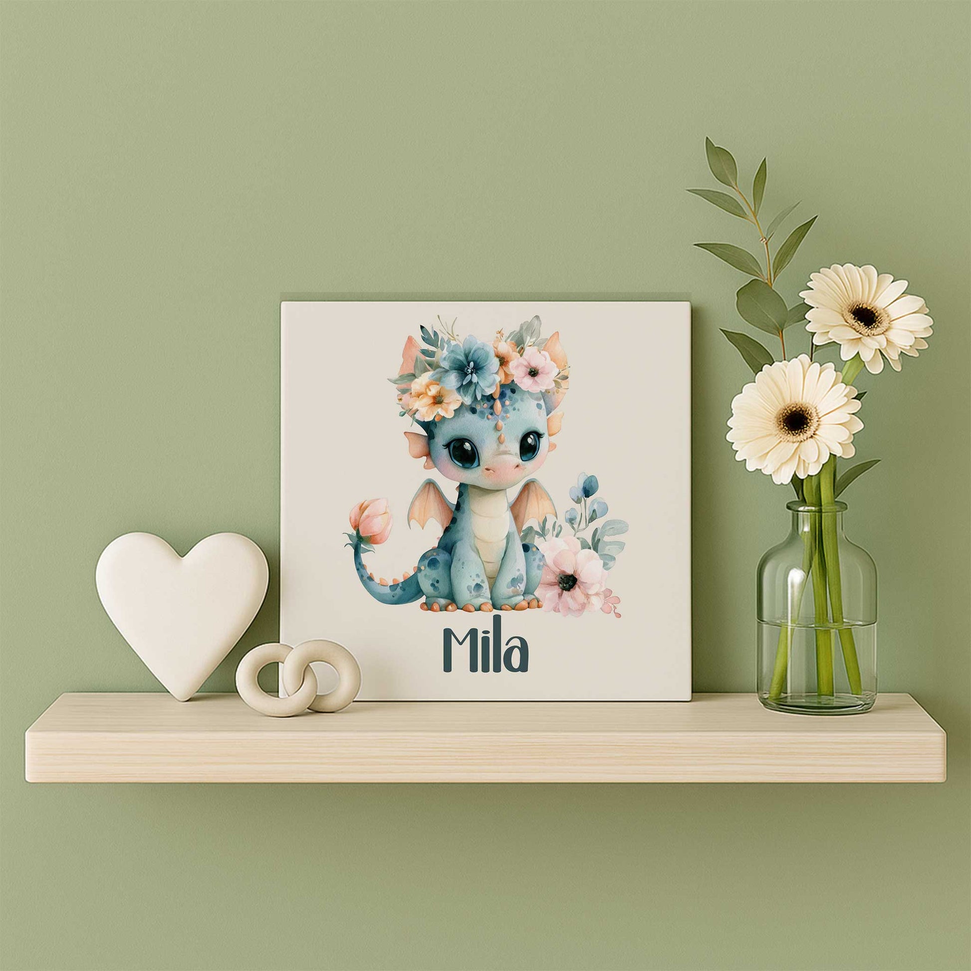 Mat keramisch tegeltje met een blauw draakje met bloemenkroon en de naam Mila op een houten wandplank naast een wit hart en een glazen vaasje met witte bloemen tegen een zachtgroene muur.