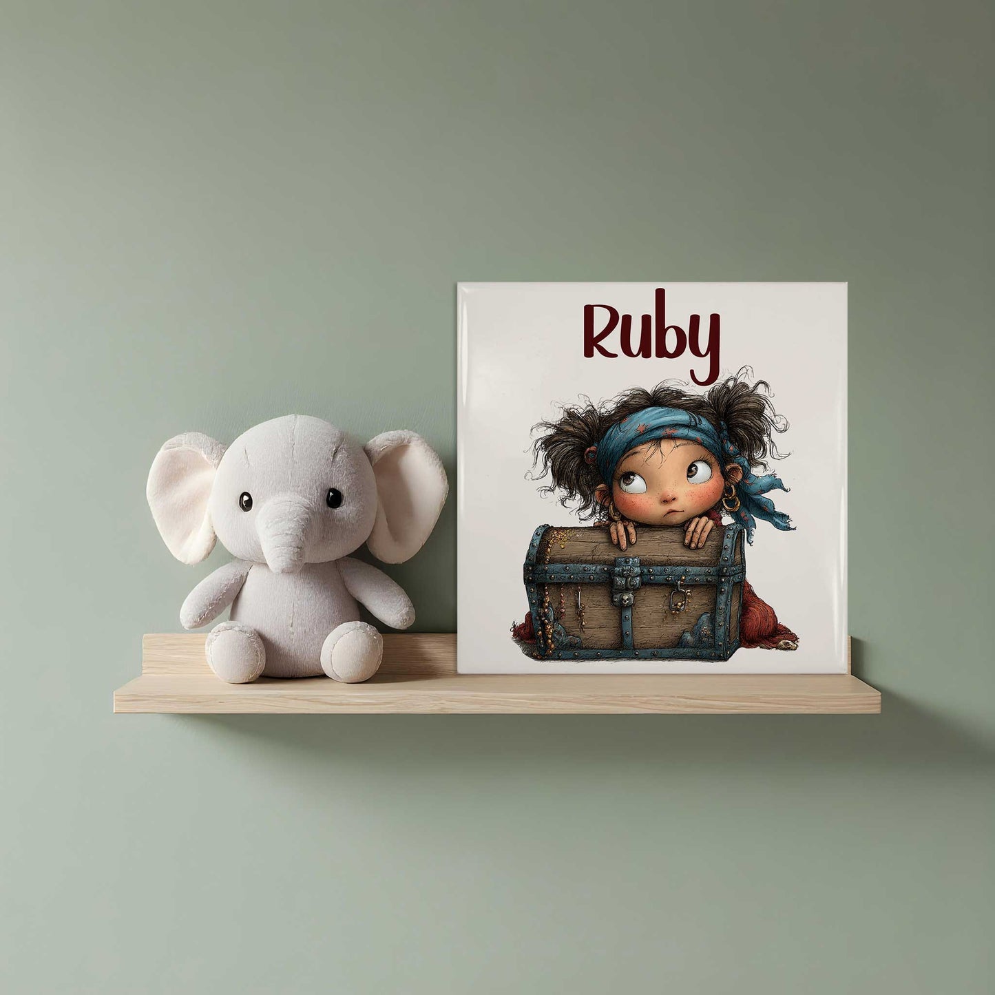 Keramisch tegeltje met de naam Ruby en een illustratie van een klein piratenmeisje dat achter een houten schatkist vandaan kijkt. Het tegeltje staat op een houten plank naast een witte knuffelolifant.