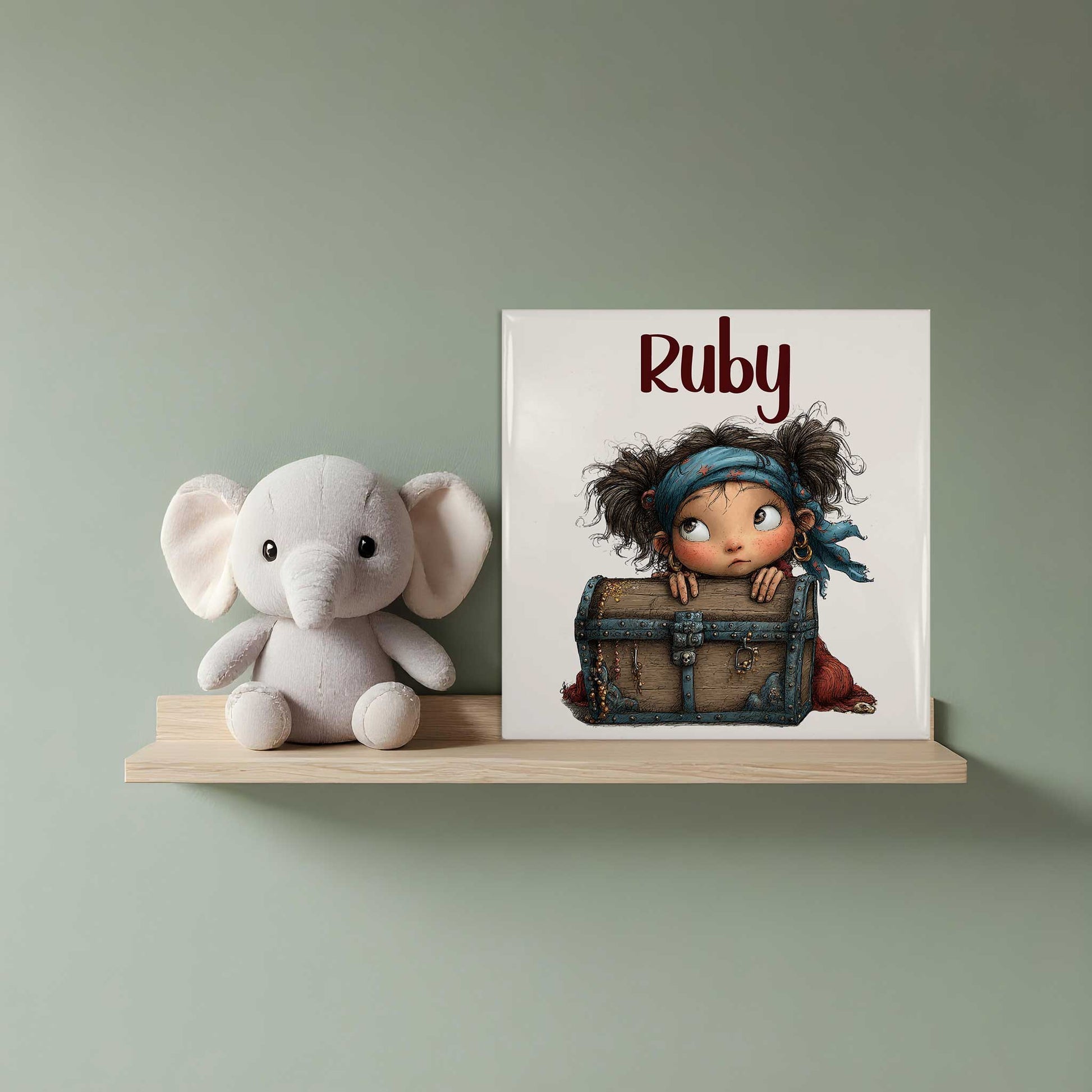 Keramisch tegeltje met de naam Ruby en een illustratie van een klein piratenmeisje dat achter een houten schatkist vandaan kijkt. Het tegeltje staat op een houten plank naast een witte knuffelolifant.