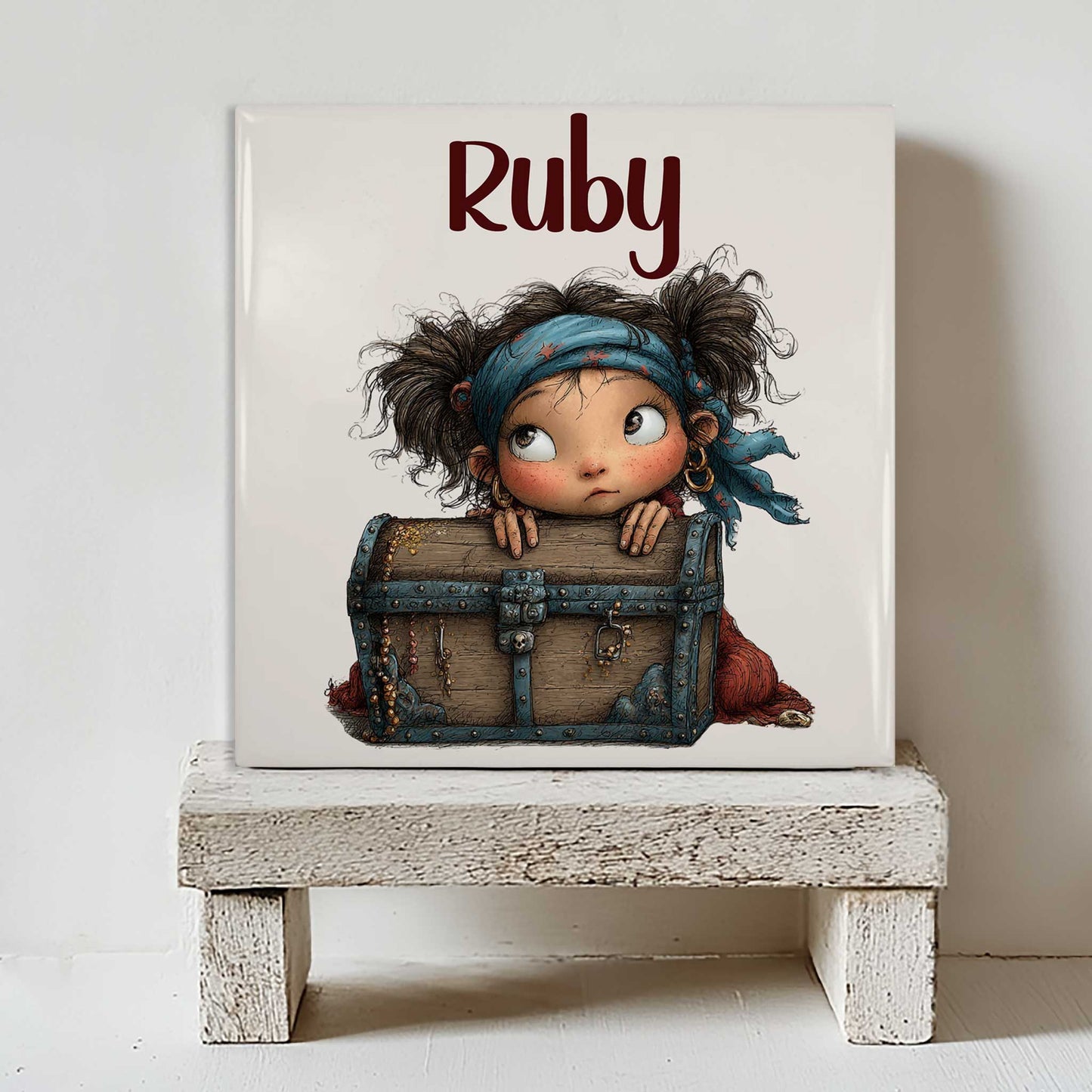 Keramisch tegeltje met de naam Ruby en een illustratie van een klein piratenmeisje achter een houten schatkist. Het tegeltje staat op een wit houten krukje tegen een lichte muur.
