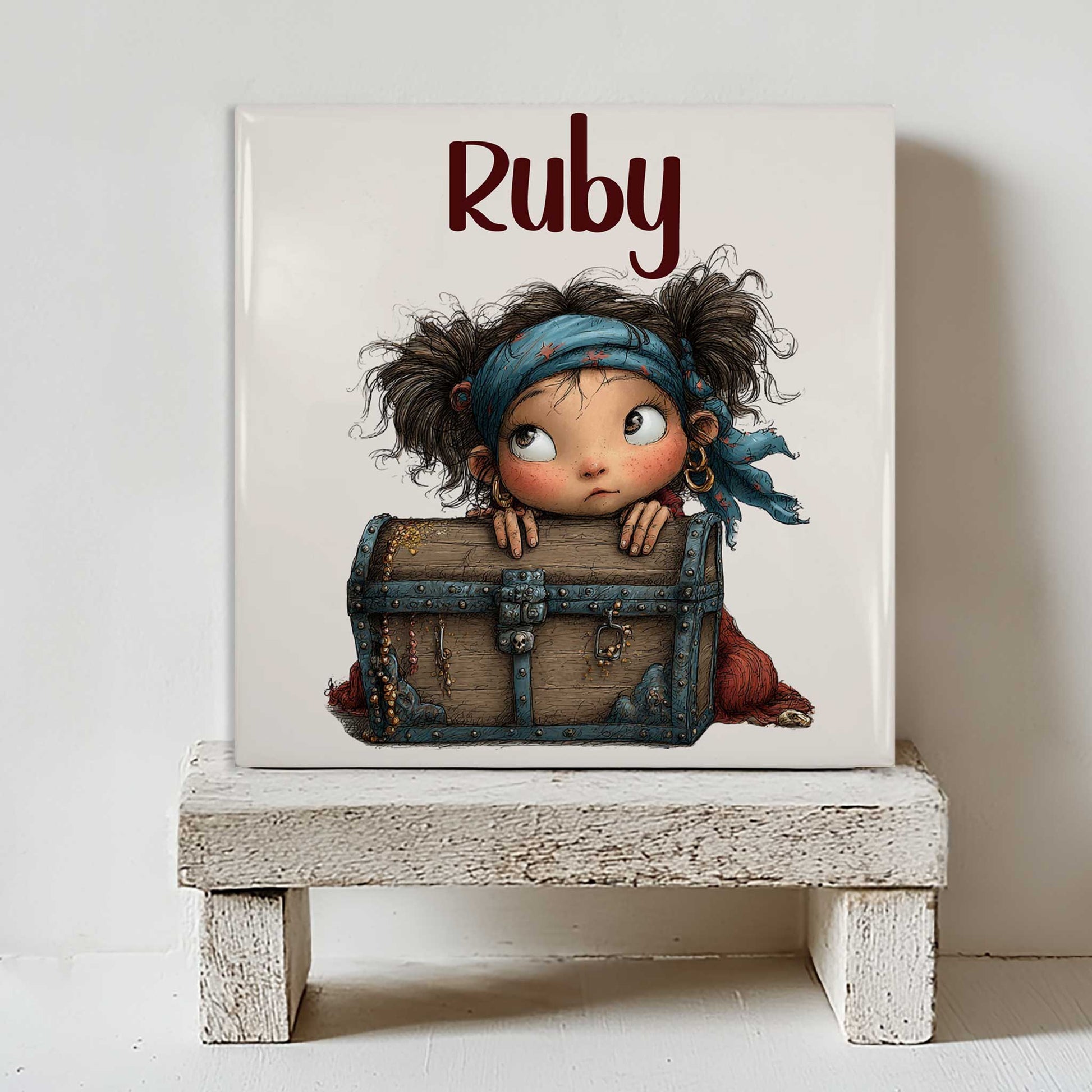 Keramisch tegeltje met de naam Ruby en een illustratie van een klein piratenmeisje achter een houten schatkist. Het tegeltje staat op een wit houten krukje tegen een lichte muur.