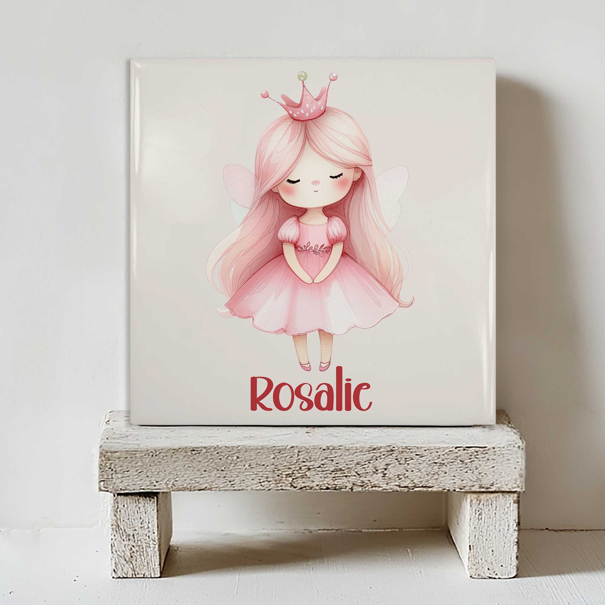 Keramisch tegeltje met een schattige prinses met lang roze haar, een roze kroontje en kleine vleugeltjes. Onder de illustratie staat de naam Rosalie. Het tegeltje staat op een wit houten krukje.
