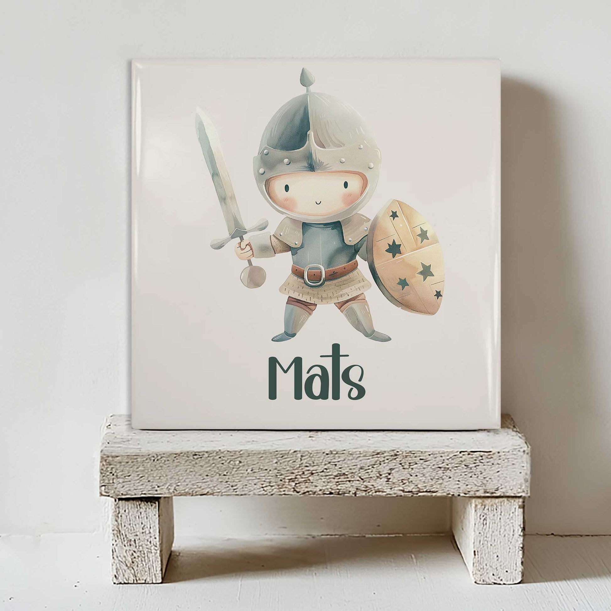 Keramisch tegeltje met een lieve ridder die een zwaard en een schild vasthoudt, gepersonaliseerd met de naam Mats, staand op een wit houten krukje.