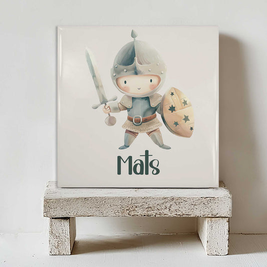 Keramisch tegeltje met een lieve ridder die een zwaard en een schild vasthoudt, gepersonaliseerd met de naam Mats, staand op een wit houten krukje.