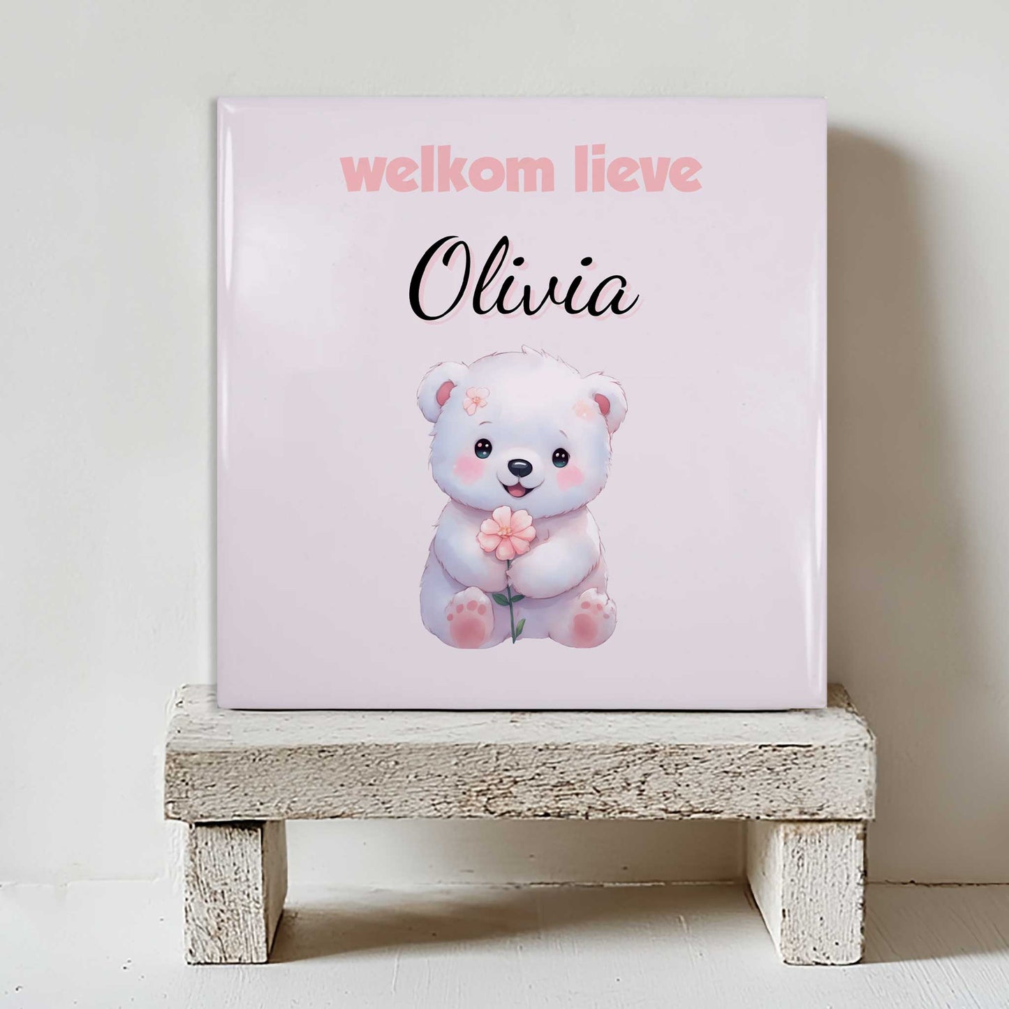 Keramisch tegeltje met een illustratie van een beer met een bloem en de tekst “welkom lieve Olivia”, geplaatst op een witte houten kruk