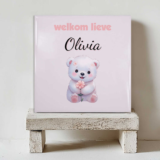 Keramisch tegeltje met een illustratie van een beer met een bloem en de tekst “welkom lieve Olivia”, geplaatst op een witte houten kruk