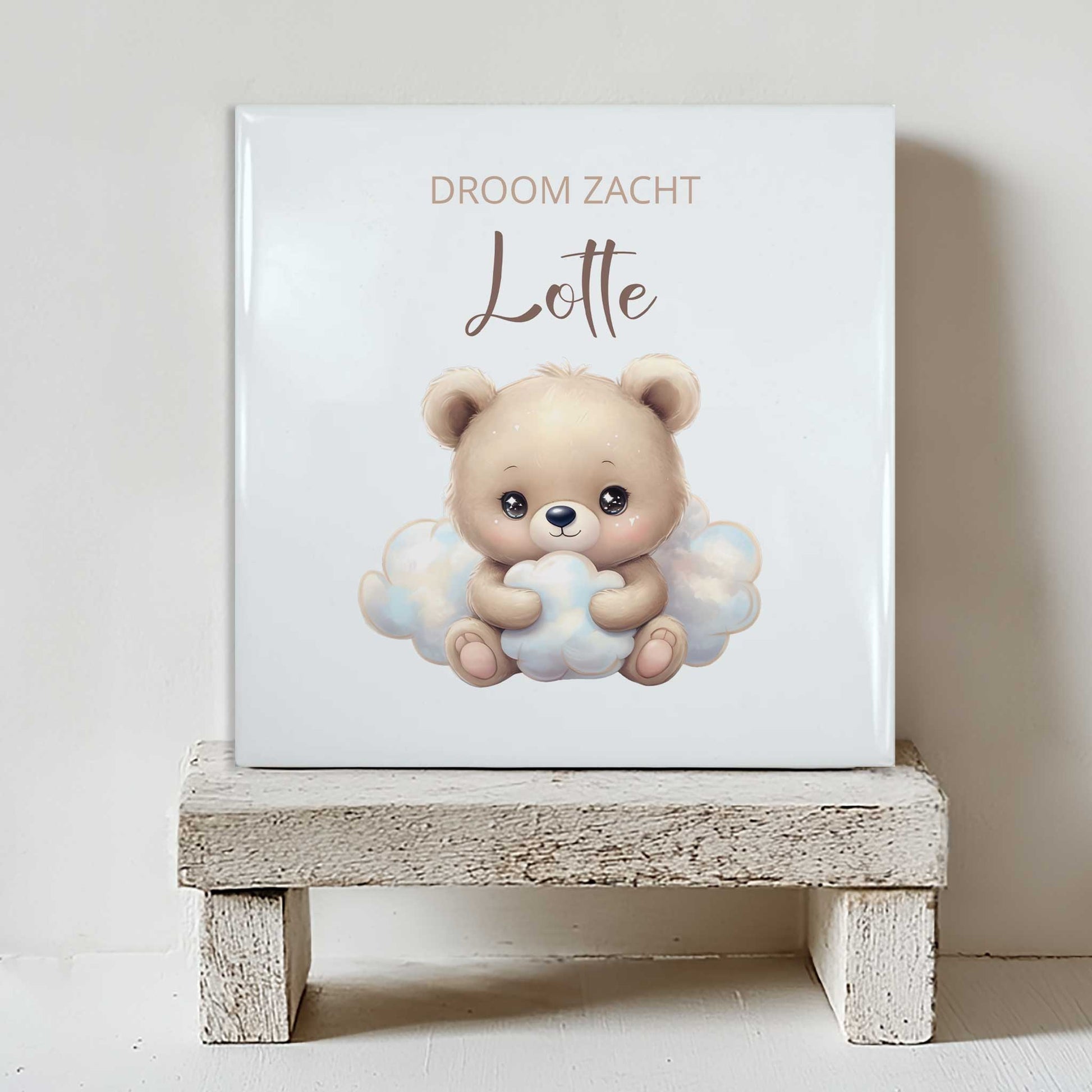 Keramisch tegeltje met een illustratie van een beer op een wolk en de tekst “droom zacht Lotte” in beige tinten, geplaatst op een witte houten kruk