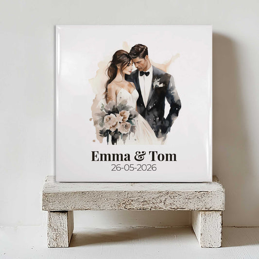 Keramisch tegeltje met een illustratie van een bruidspaar en de namen Emma & Tom met de datum 26-05-2026, geplaatst op een witte houten kruk
