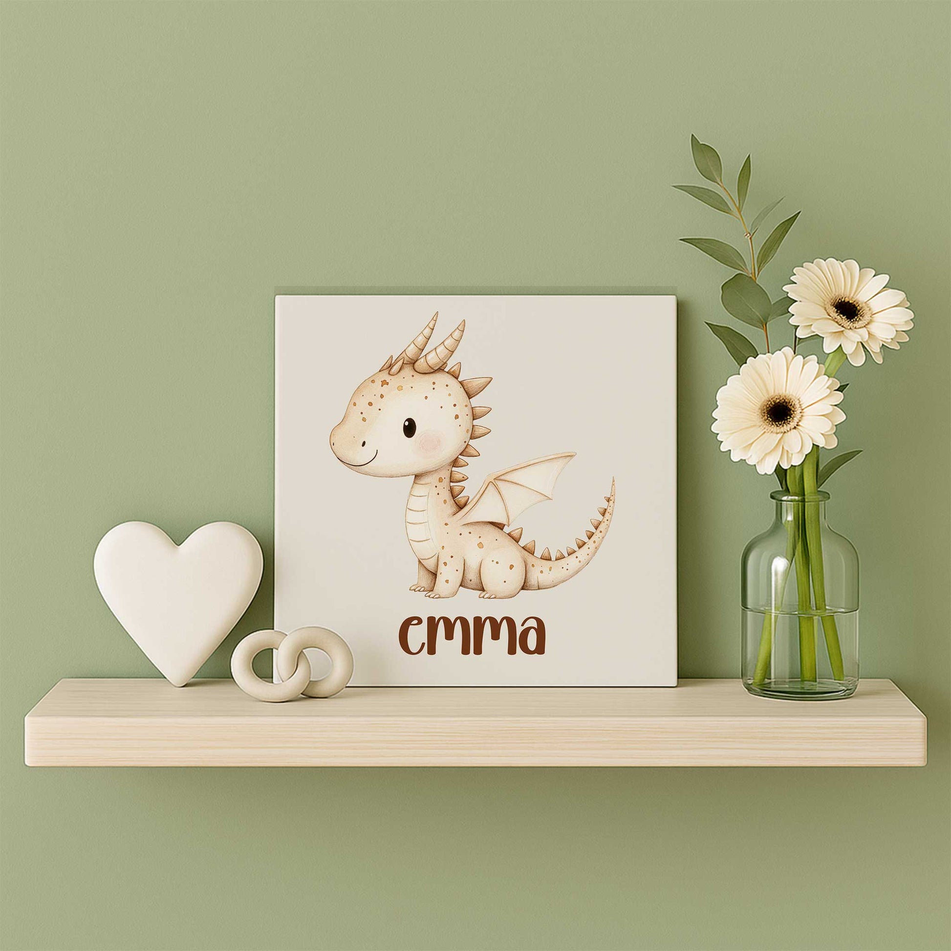 Mat keramisch tegeltje met een beige draakje en de naam Emma op een houten wandplank naast een wit hart en een glazen vaasje met witte bloemen tegen een zachtgroene muur.