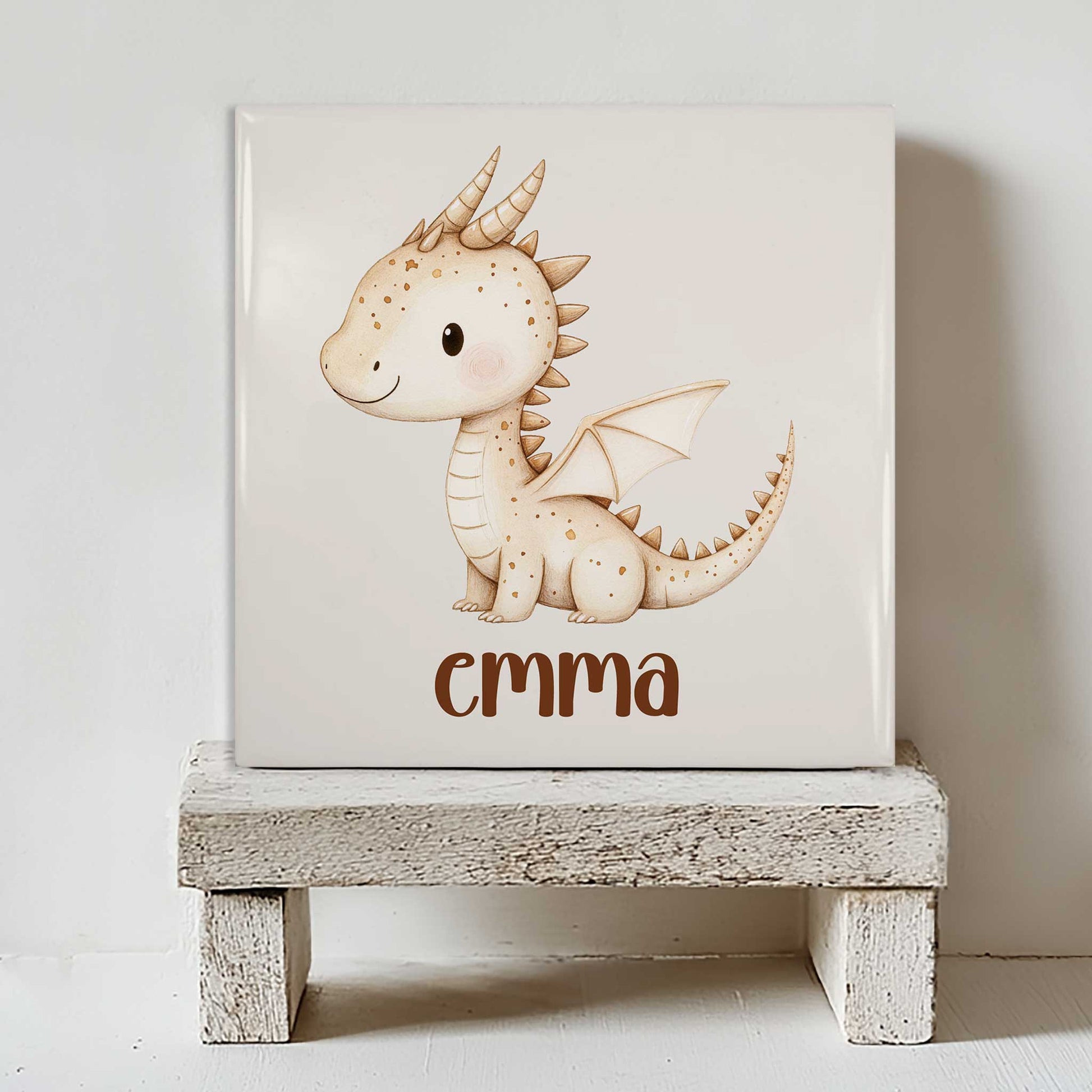Mat keramisch tegeltje met een beige draakje met kleine vleugels en de gepersonaliseerde naam Emma, staand op een wit houten krukje tegen een lichte achtergrond.