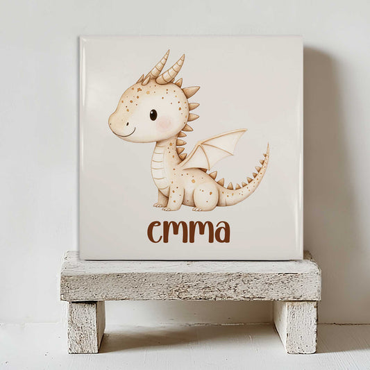 Mat keramisch tegeltje met een beige draakje met kleine vleugels en de gepersonaliseerde naam Emma, staand op een wit houten krukje tegen een lichte achtergrond.