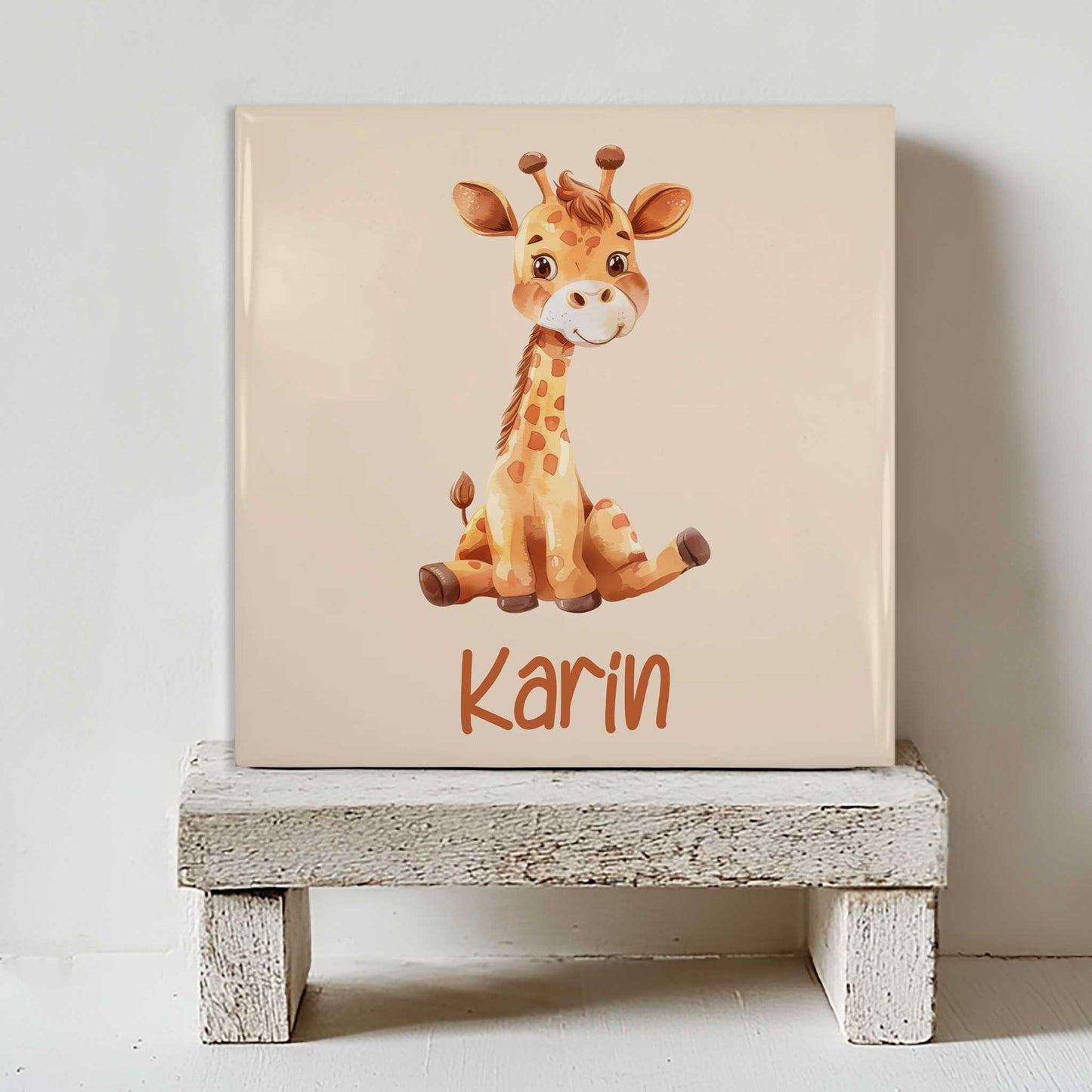 Keramisch tegeltje met een illustratie van een giraf en de naam Karin in beige tinten, geplaatst op een witte houten kruk