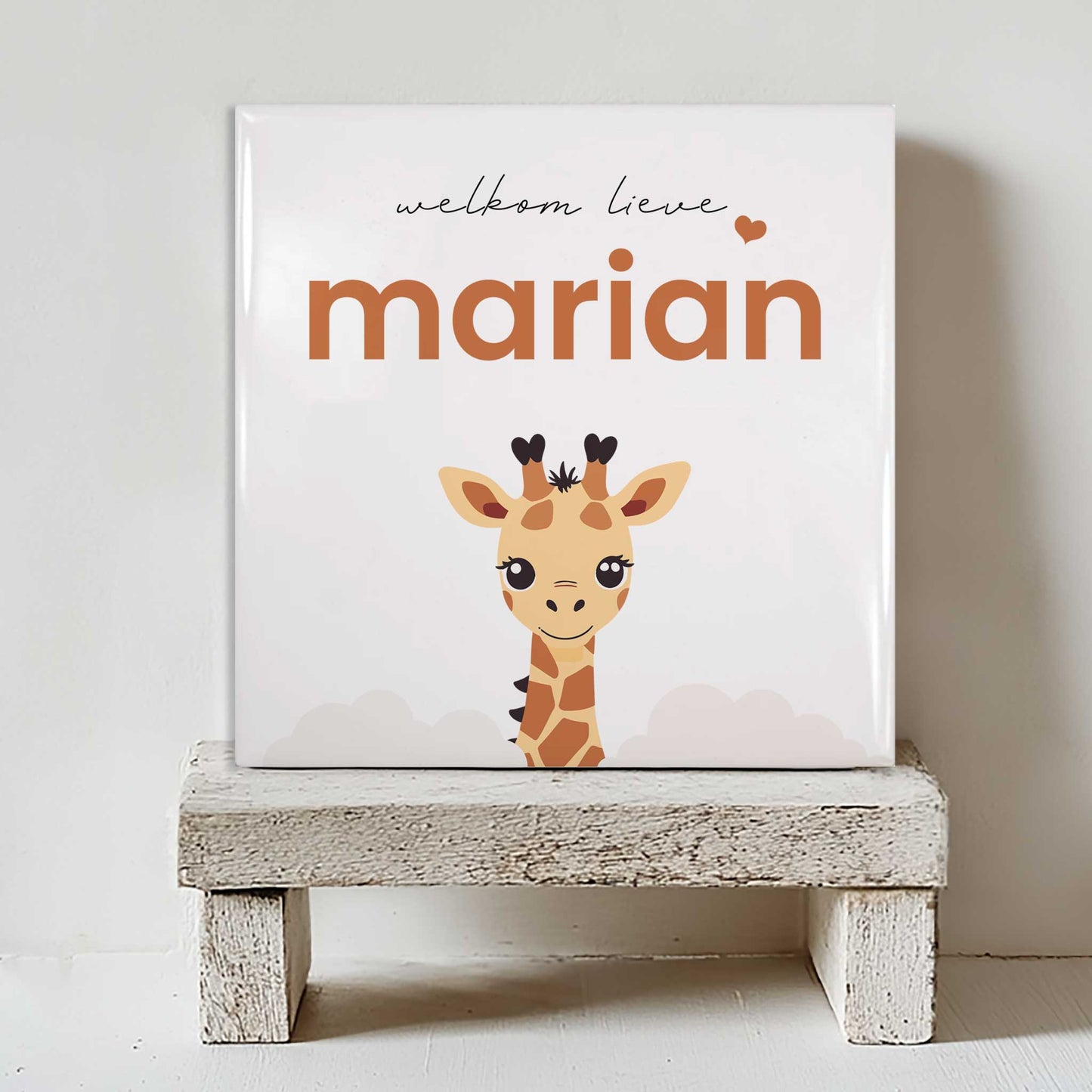 Keramisch tegeltje met een illustratie van een giraf en de tekst “welkom lieve marian”, geplaatst op een witte houten kruk