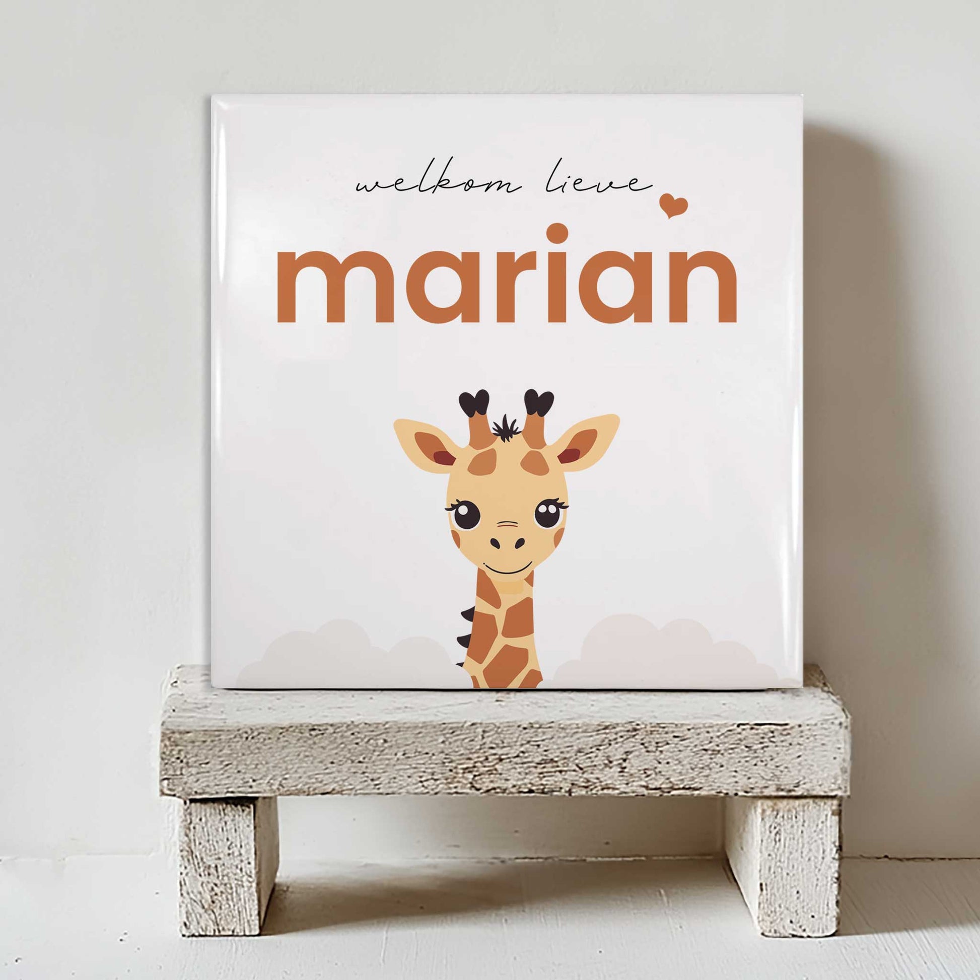 Keramisch tegeltje met een illustratie van een giraf en de tekst “welkom lieve marian”, geplaatst op een witte houten kruk