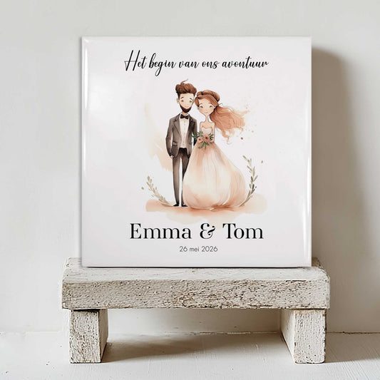 Keramisch tegeltje met de tekst “Het begin van ons avontuur” en een illustratie van een bruidspaar, met de namen Emma & Tom en de datum 26 mei 2026, geplaatst op een witte houten kruk