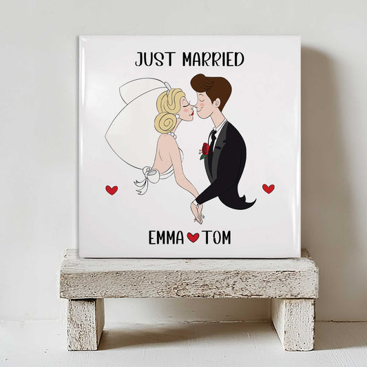 Keramisch tegeltje met de tekst “Just Married” en een illustratie van een bruidspaar, met de namen Emma en Tom en rode hartjes, geplaatst op een witte houten kruk
