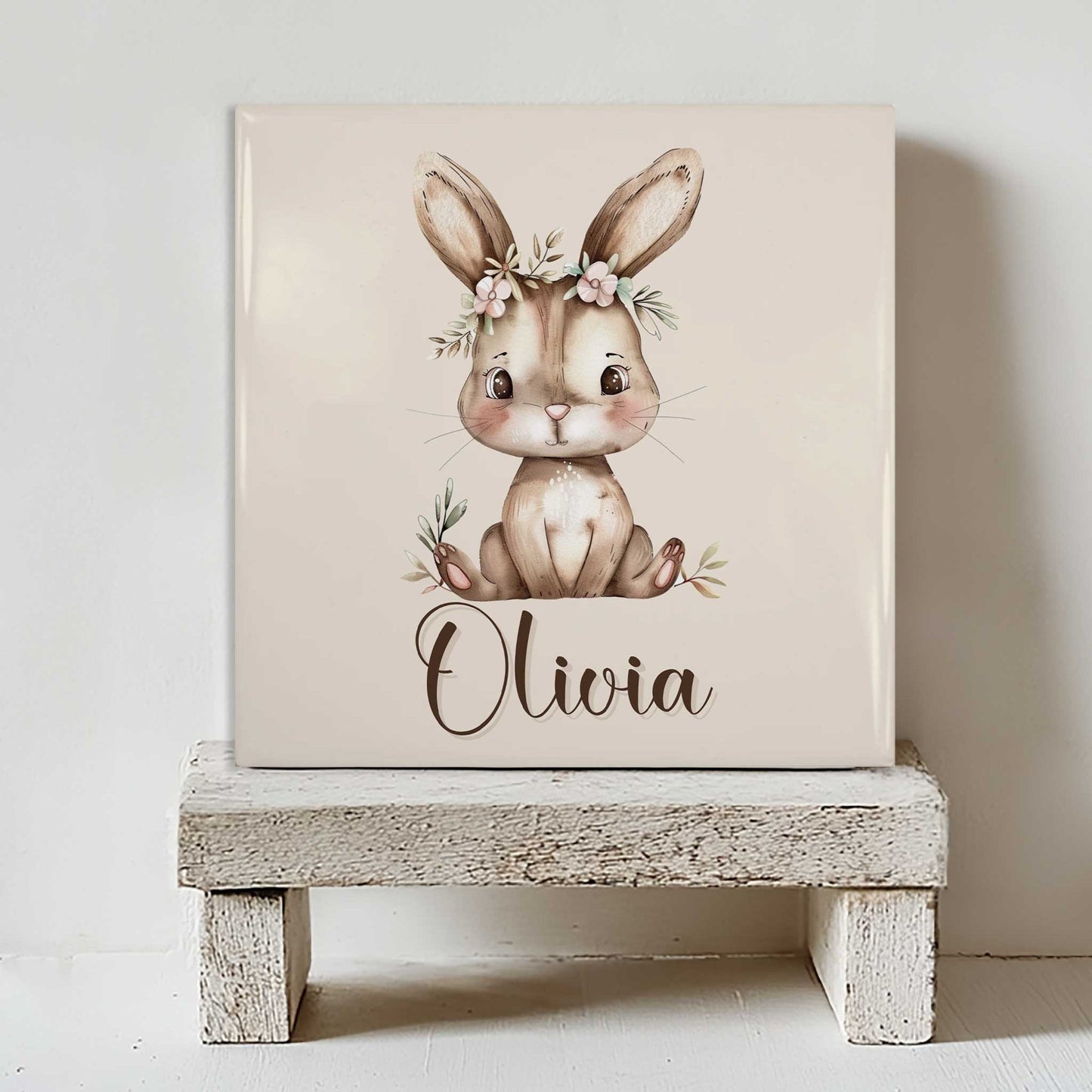 Keramisch tegeltje met een illustratie van een konijn met een bloemenkrans en de naam Olivia in beige tinten, geplaatst op een witte houten kruk