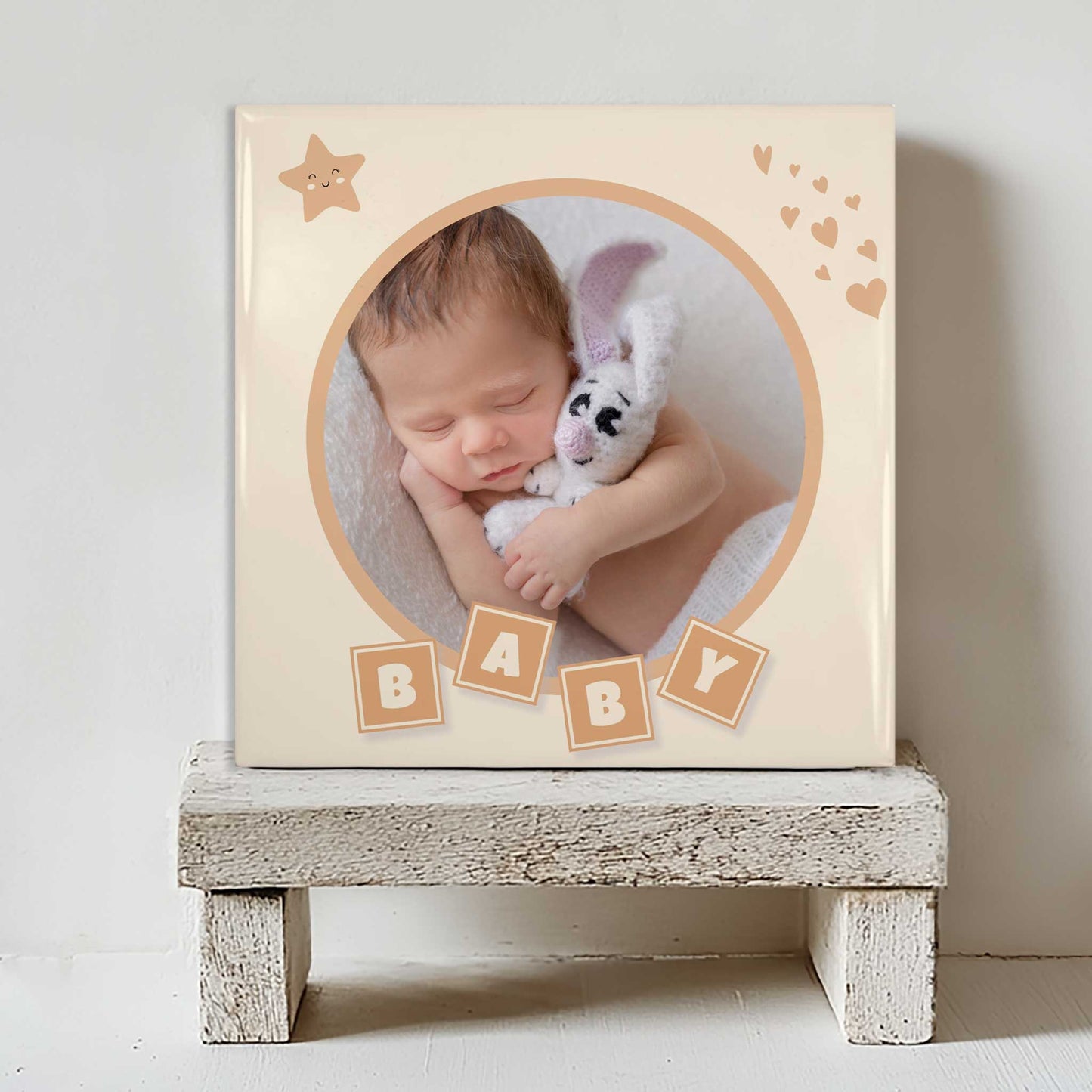 Keramisch tegeltje met een foto van een slapende baby in een rond kader, met blokjes waarop “BABY” staat en kleine hartjes, geplaatst op een witte houten kruk