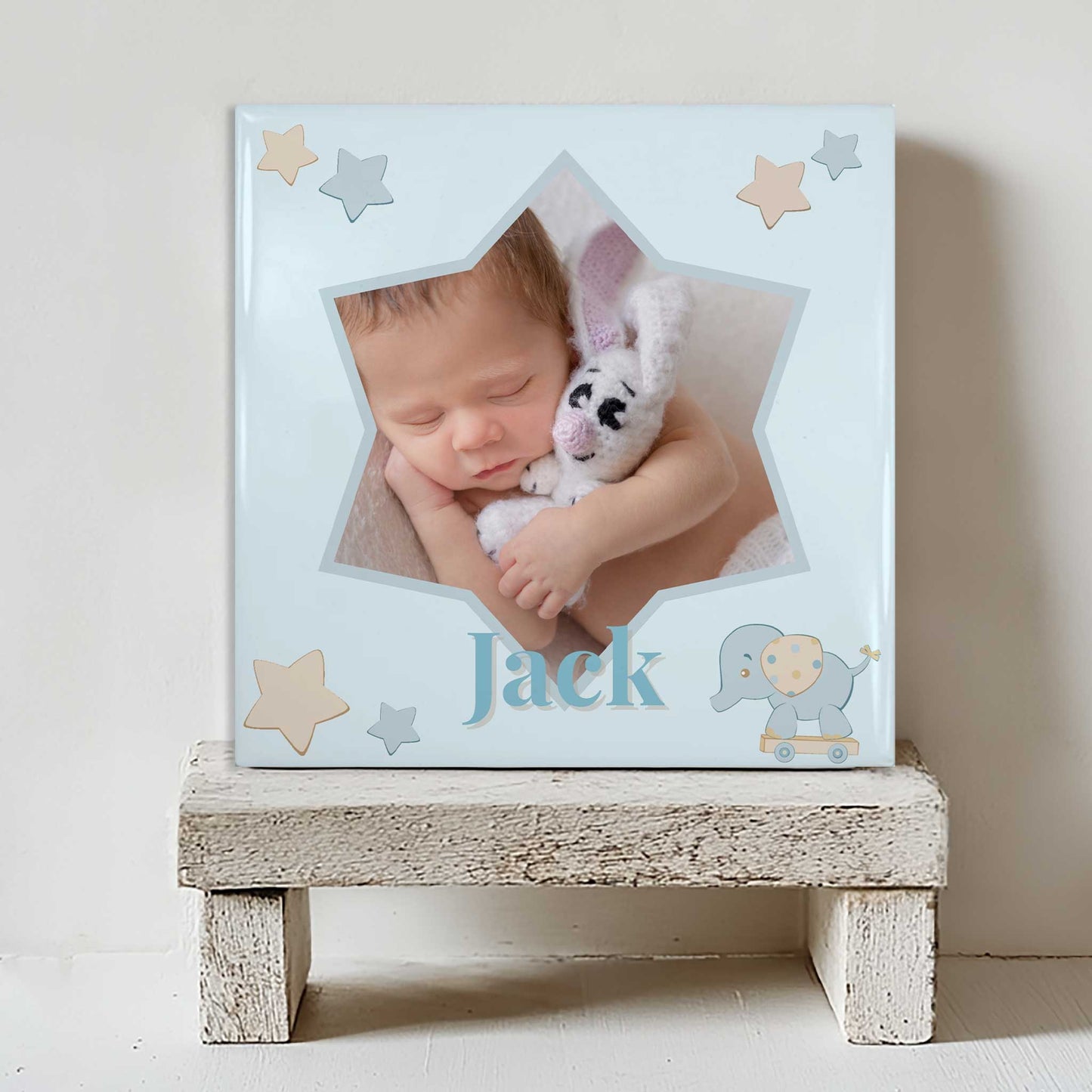 Keramisch tegeltje met een foto van een slapende baby in een stervormig kader, met de naam Jack in blauwe tinten en kleine sterren, geplaatst op een witte houten kruk