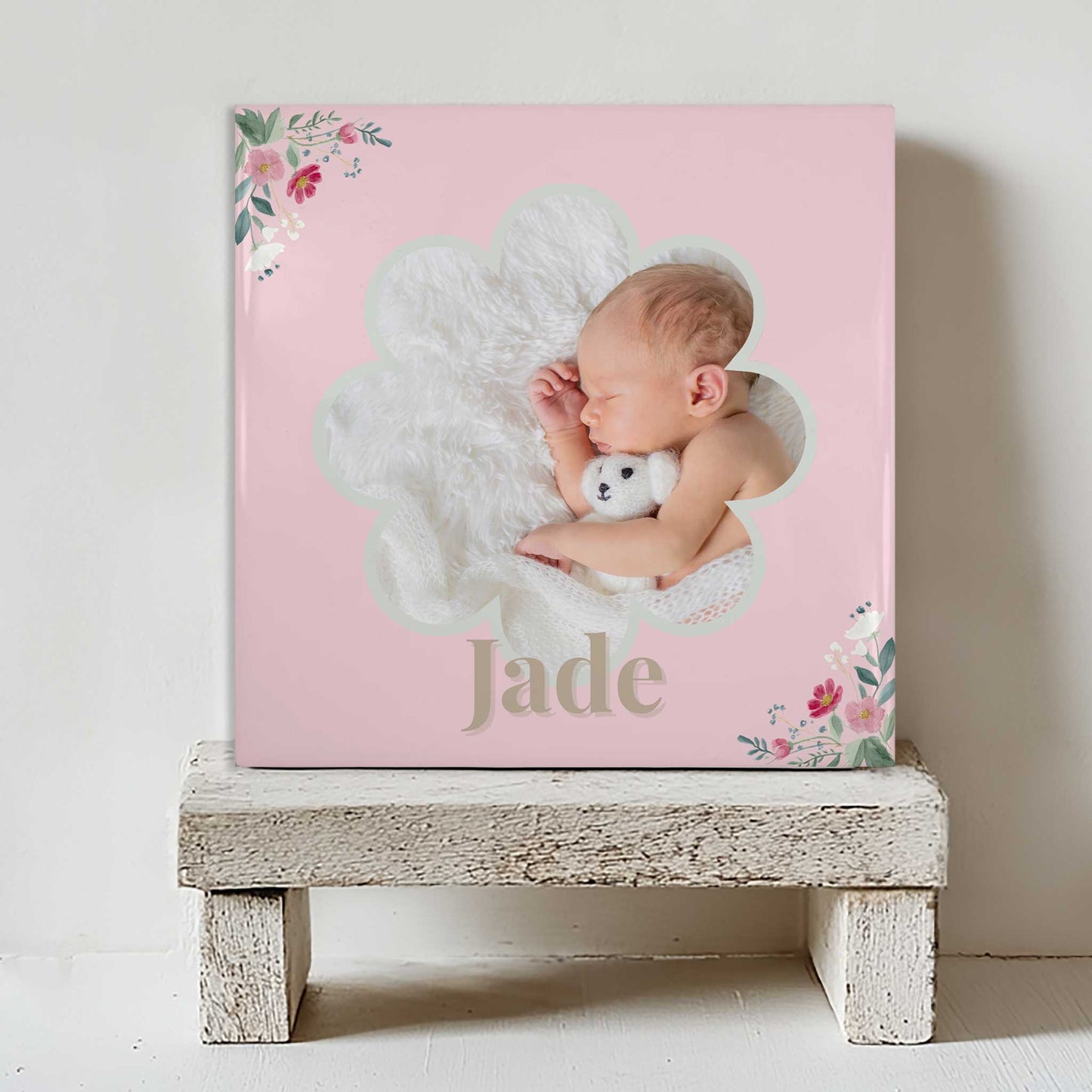 Keramisch tegeltje met een foto van een slapende baby in een bloemvormig kader, met de naam Jade in roze tinten en bloemen in de hoeken, geplaatst op een witte houten kruk