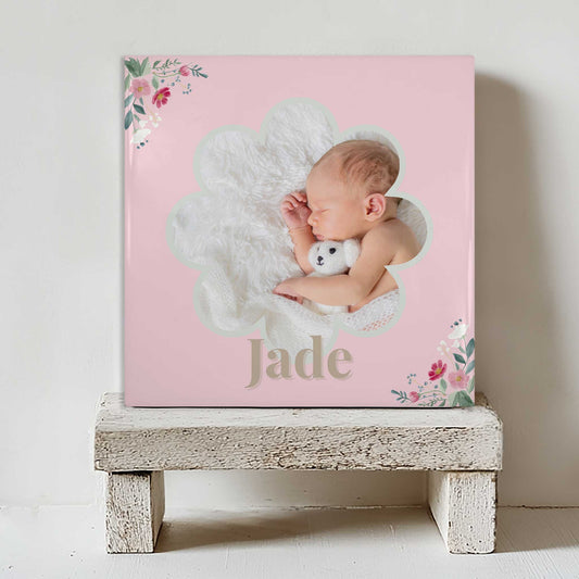 Keramisch tegeltje met een foto van een slapende baby in een bloemvormig kader, met de naam Jade in roze tinten en bloemen in de hoeken, geplaatst op een witte houten kruk
