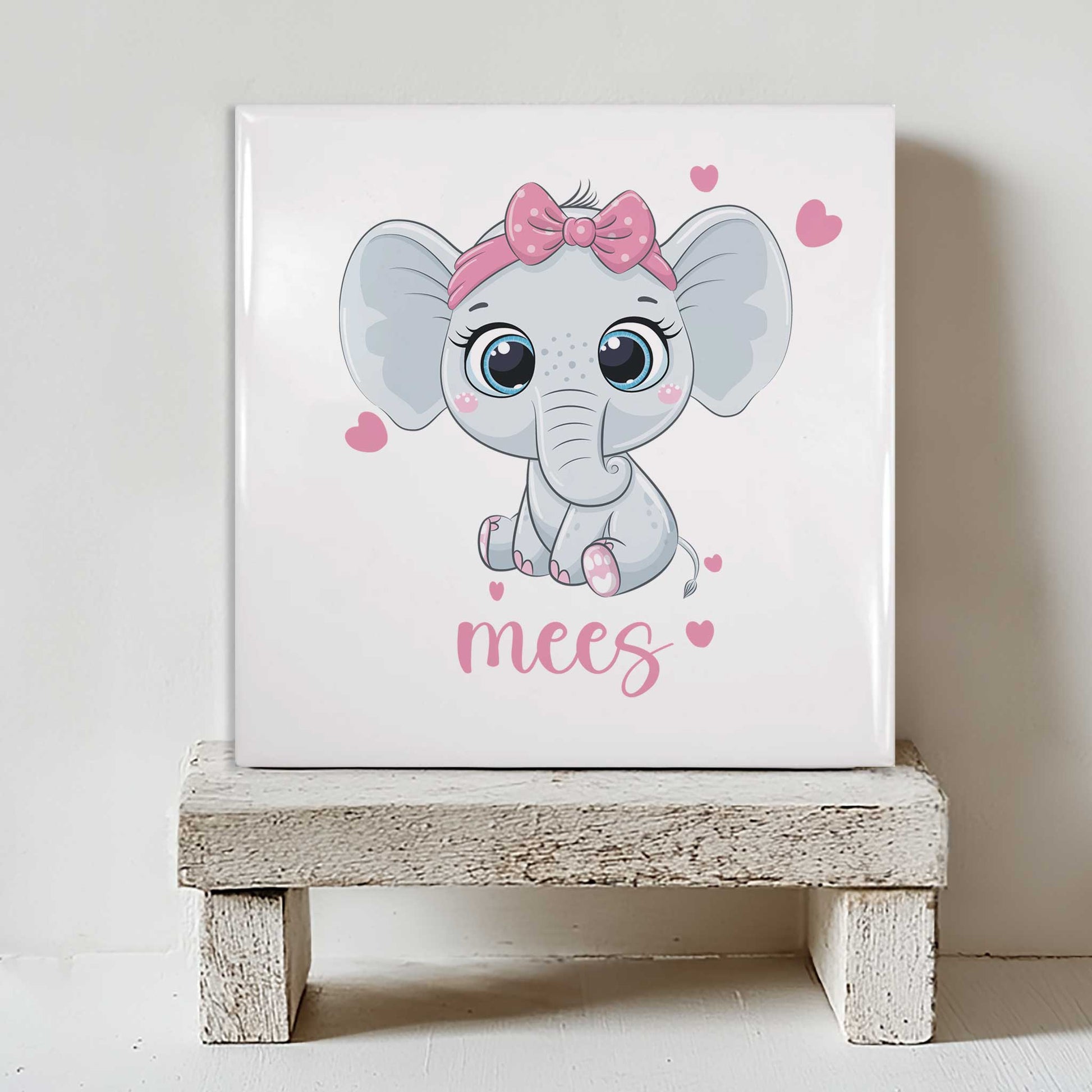 Keramisch tegeltje met een illustratie van een olifant met een strik en de naam mees in roze tinten, geplaatst op een witte houten kruk
