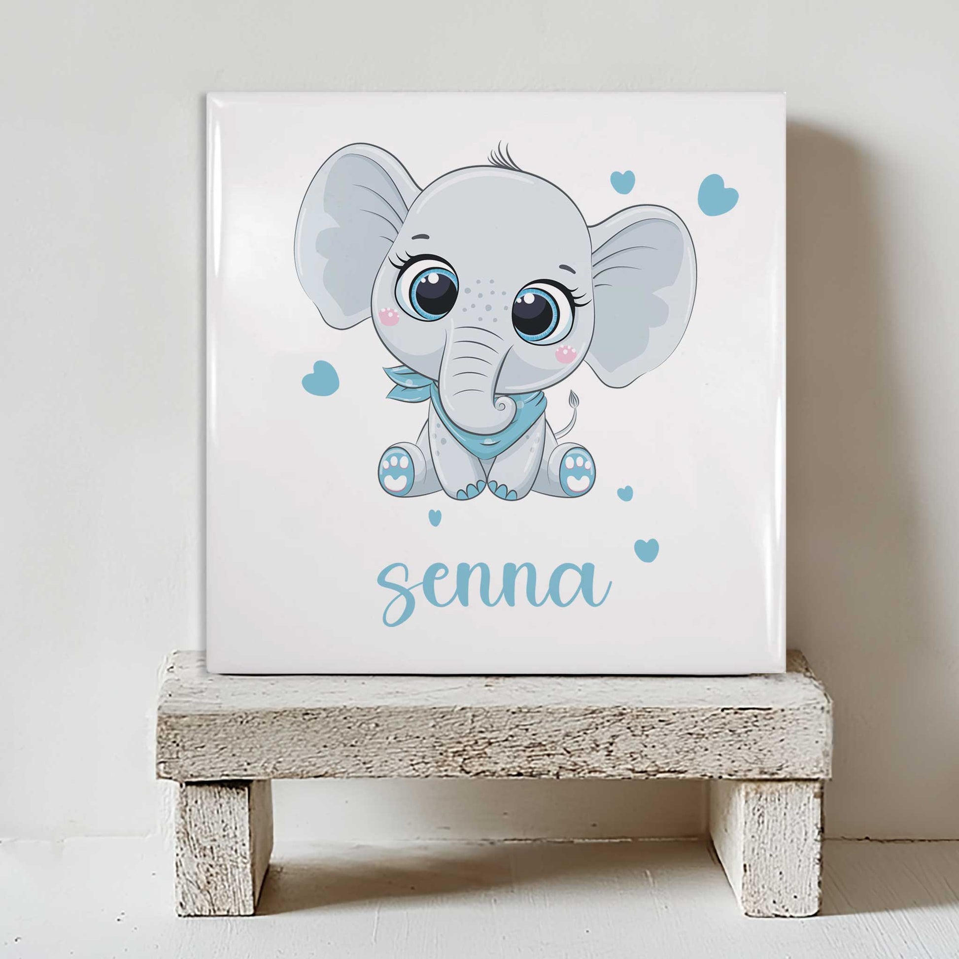 Keramisch tegeltje met een illustratie van een olifant en de naam Senna in roze tinten, geplaatst op een witte houten kruk