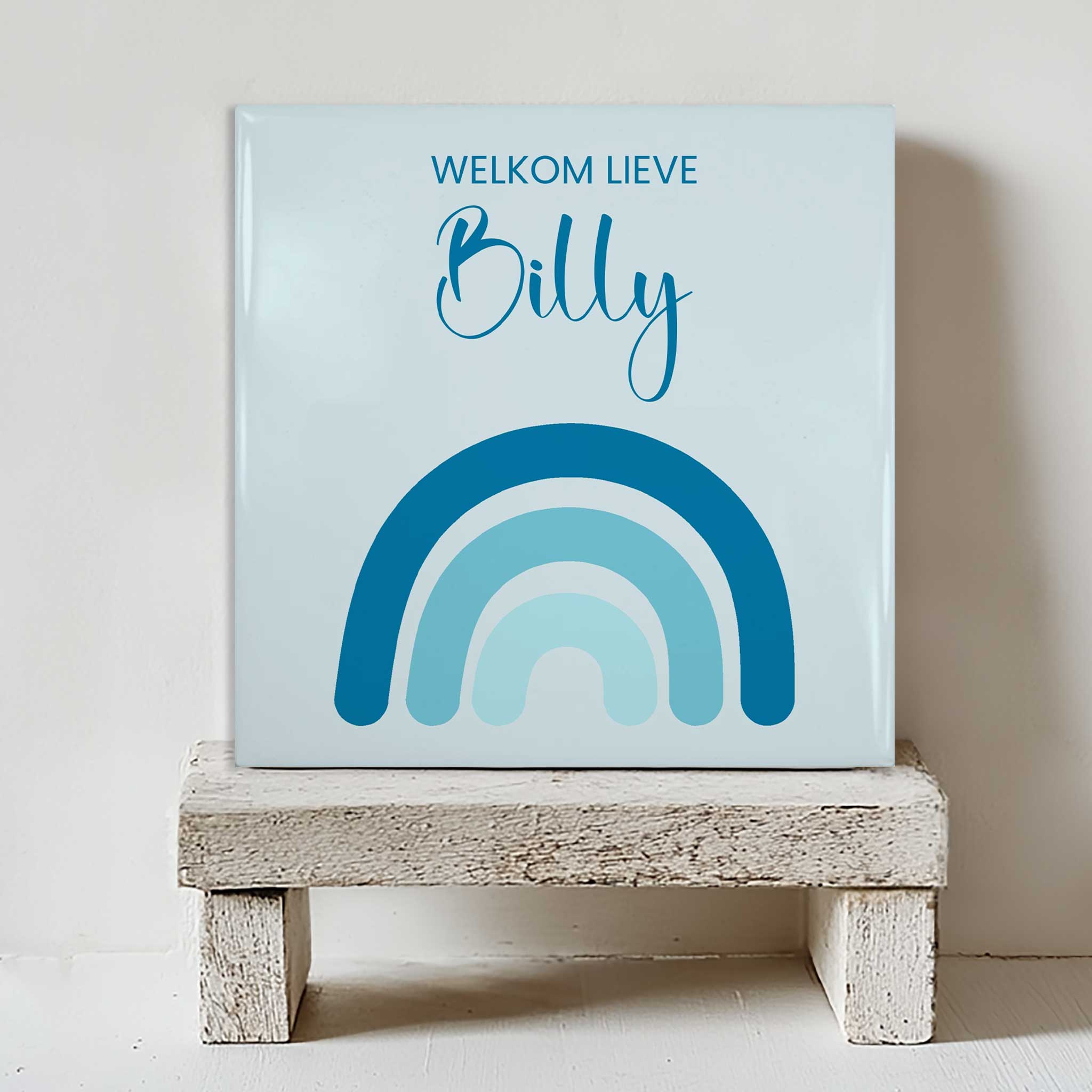 Keramisch tegeltje met een blauwe regenboog en de tekst “welkom lieve Billy”, geplaatst op een witte houten kruk