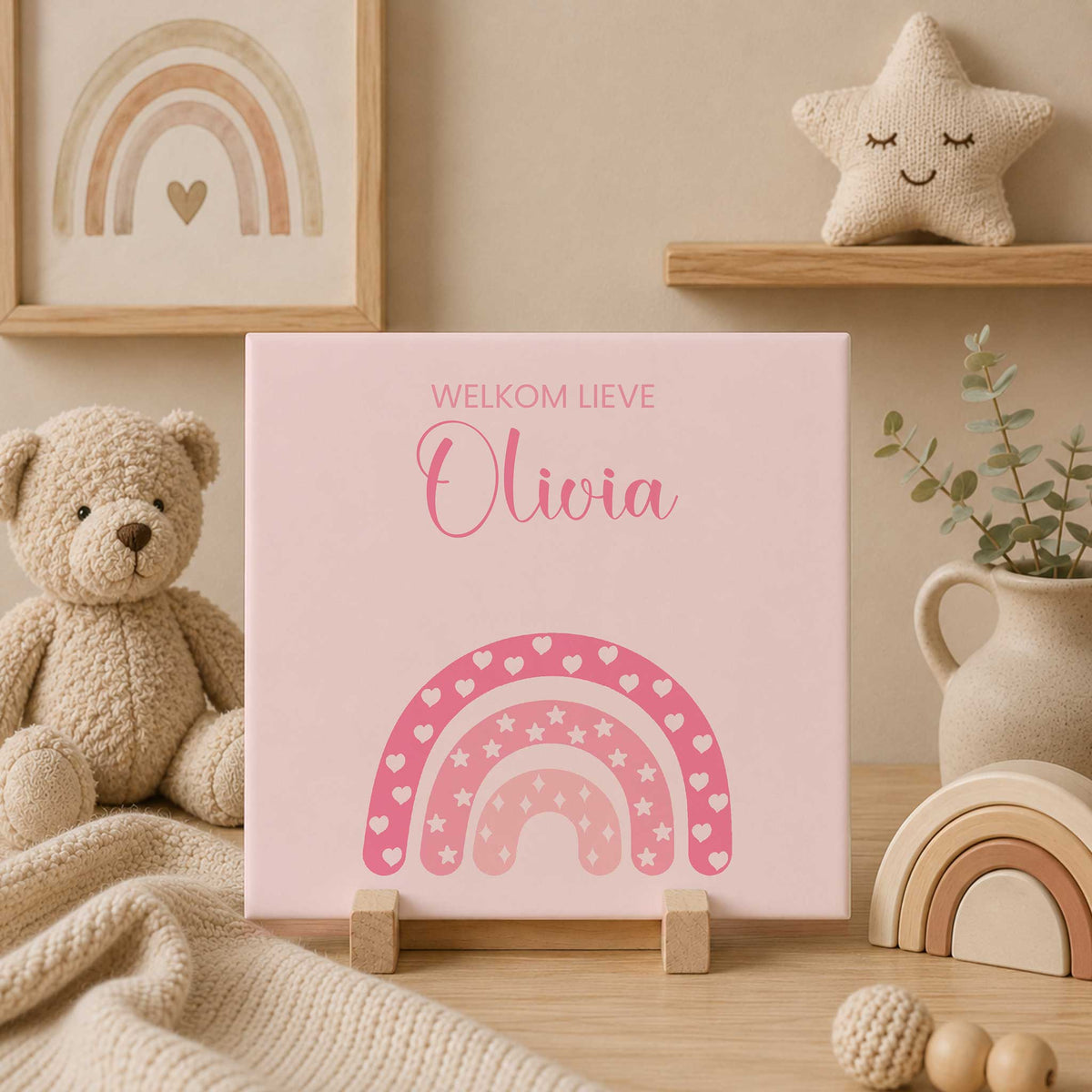 Roze gepersonaliseerd geboortetegeltje met de tekst ‘Welkom lieve Olivia’ en een regenboog met hartjes, sterren en fonkeltjes, staand op een houten standaard in een beige babykamer met knuffelbeer.