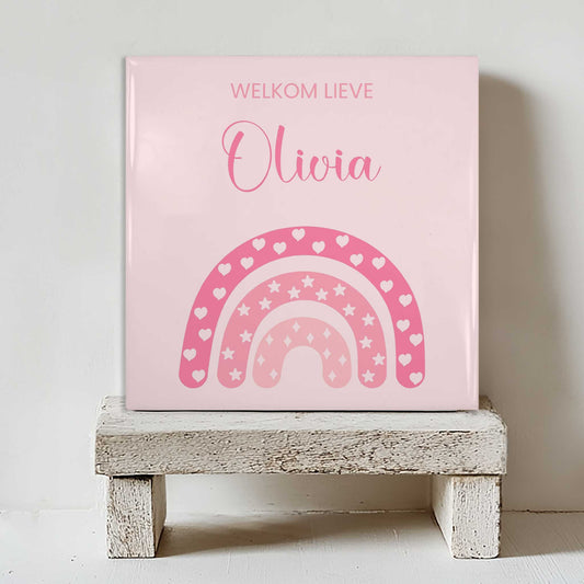 Keramisch tegeltje met een roze regenboog met hartjes en sterren en de tekst “welkom lieve Olivia”, geplaatst op een witte houten kruk