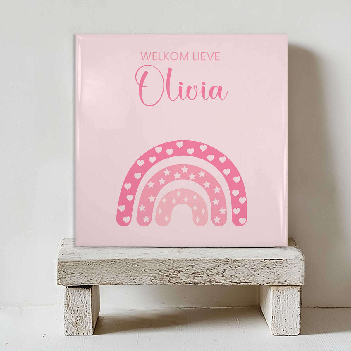 Keramisch tegeltje met een roze regenboog met hartjes en sterren en de tekst “welkom lieve Olivia”, geplaatst op een witte houten kruk
