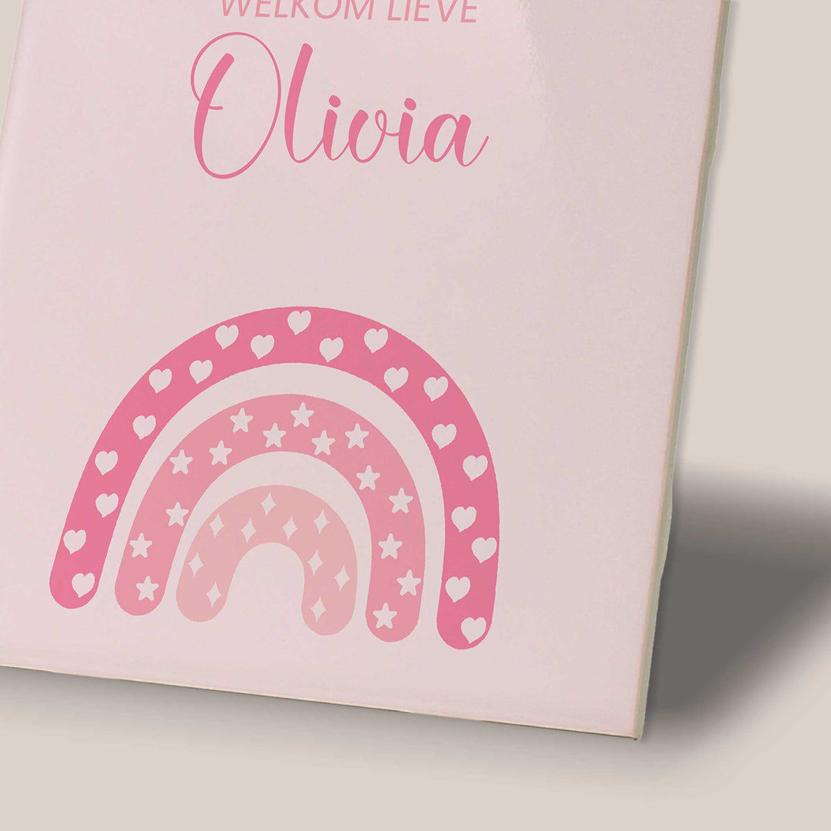 Close-up van een roze gepersonaliseerd geboortetegeltje met de tekst ‘Welkom lieve Olivia’ en een regenboog in verschillende roze tinten, versierd met hartjes, sterren en fonkeltjes.