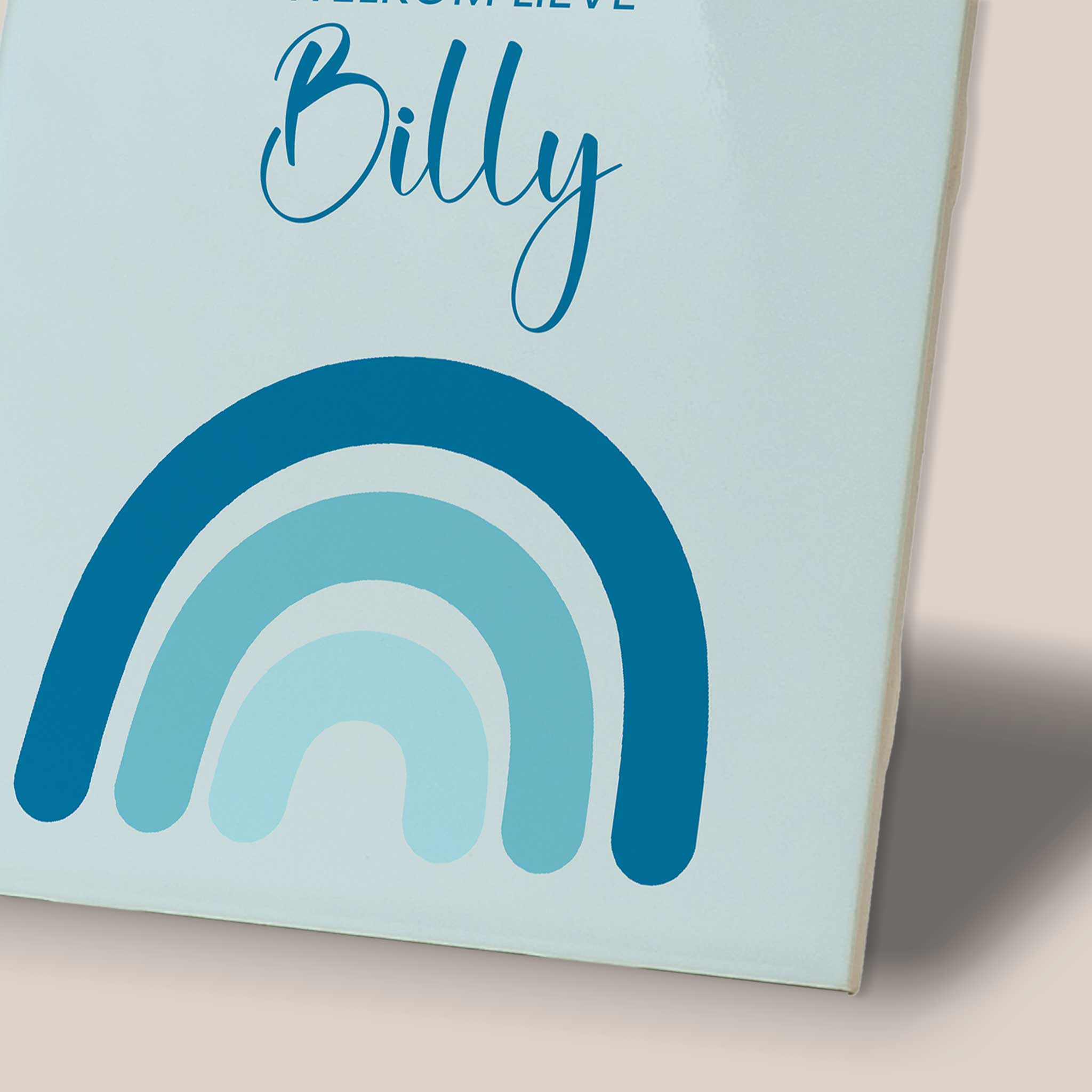 Close-up van een blauw gepersonaliseerd geboortetegeltje met de tekst ‘Welkom lieve Billy’ en een regenboog in verschillende tinten blauw.