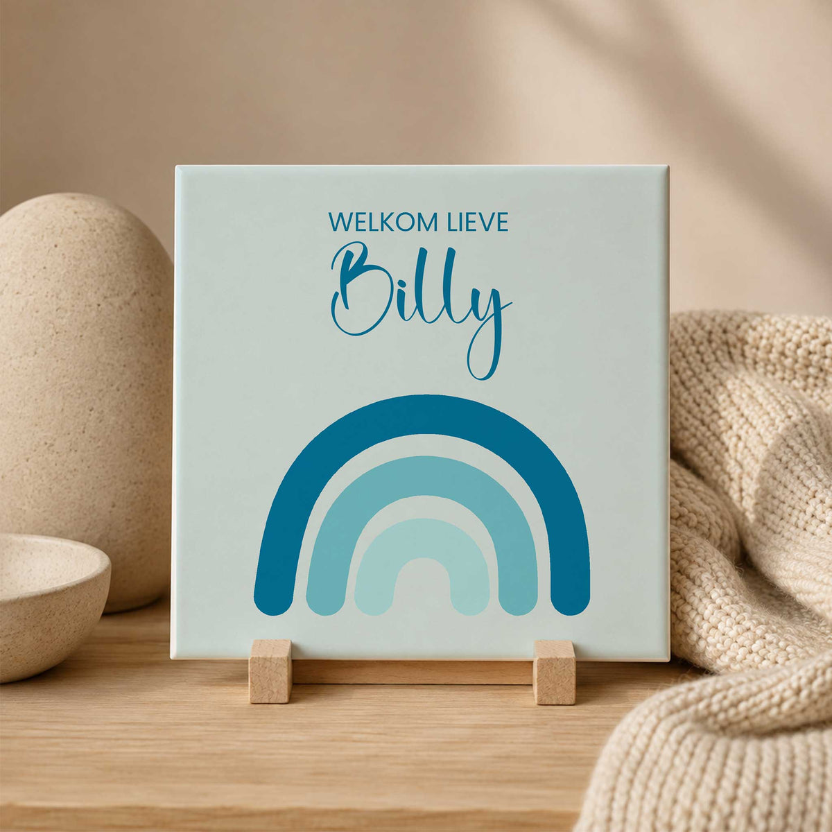 Blauw gepersonaliseerd geboortetegeltje met de tekst ‘Welkom lieve Billy’ en een regenboog in verschillende blauwtinten, staand op een houten standaard in een beige setting.