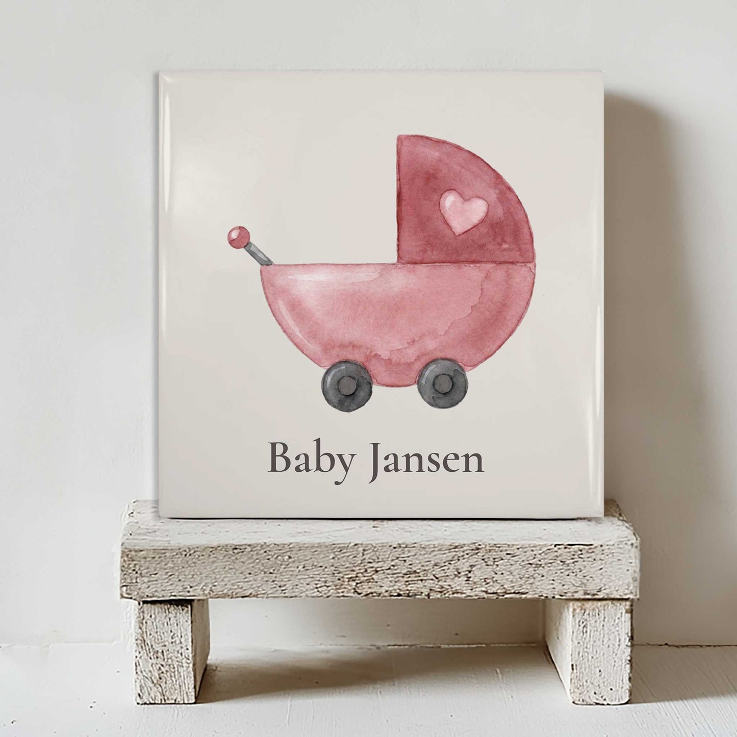 Gepersonaliseerd babytegeltje met roze watercolor kinderwagen en naam Baby Jansen geplaatst op een wit houten krukje