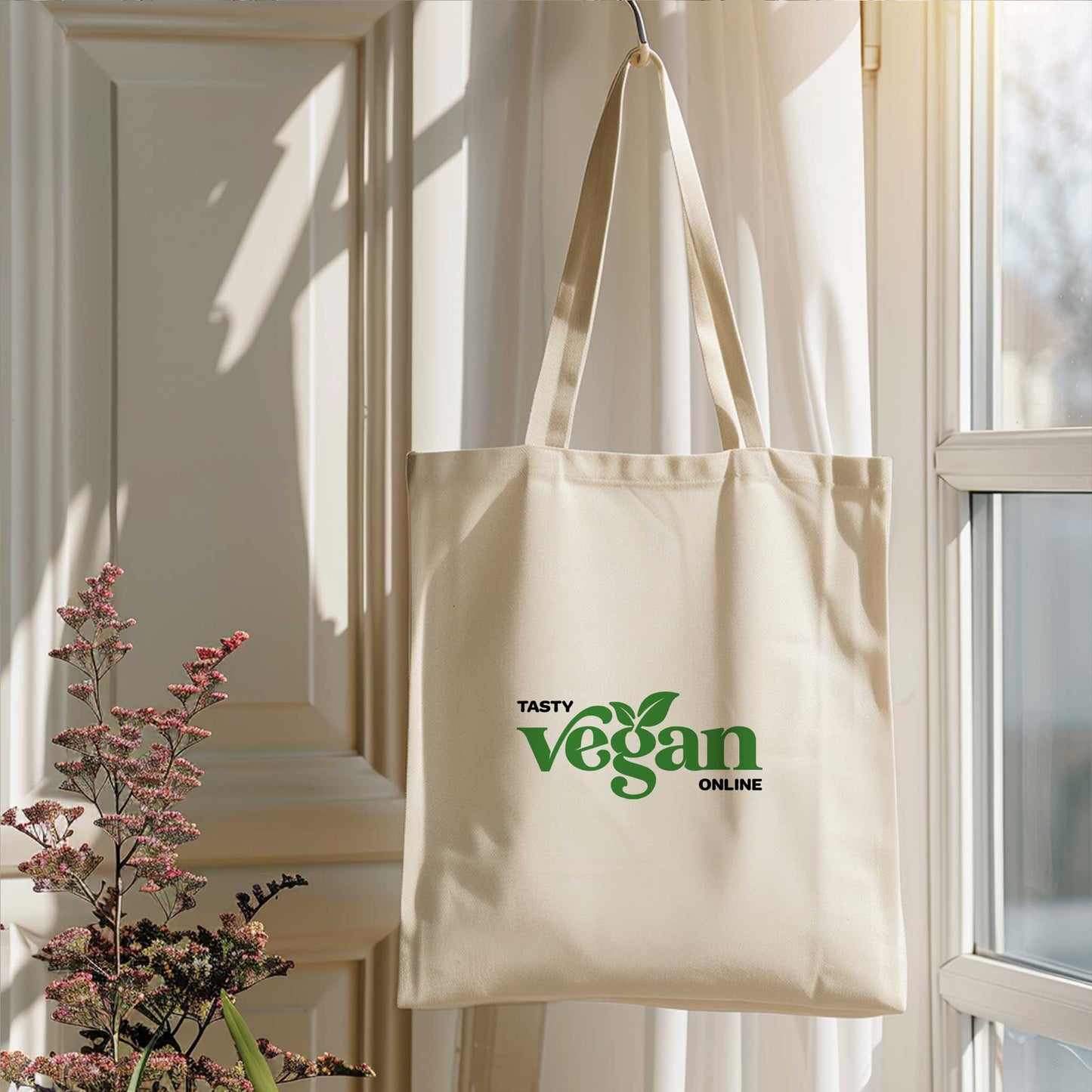 Beige totebag bedrukt met een groen bedrijfslogo, hangend naast een raam in een lichte ruimte met bloemen op de voorgrond.