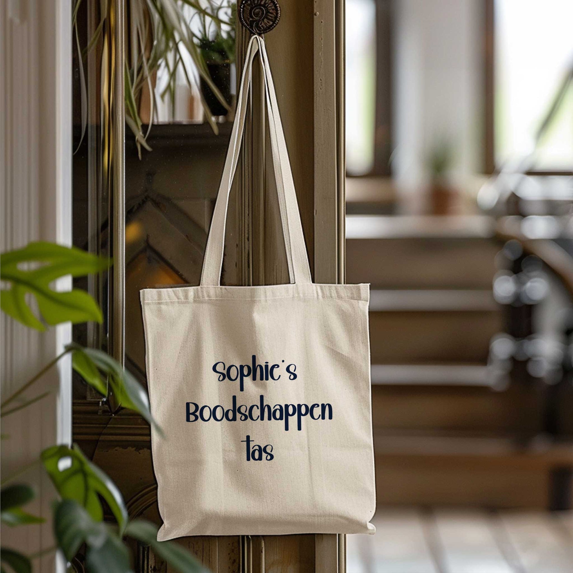 Beige totebag bedrukt met de tekst 'Sophie's Boodschappentas', hangend aan een haak in een hal met een trap op de achtergrond.