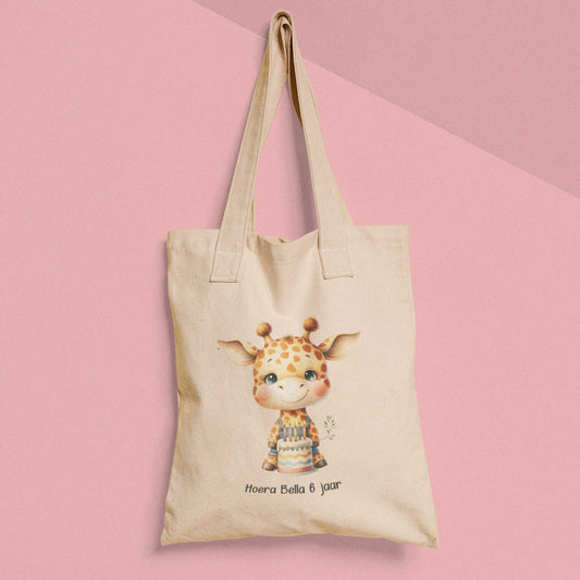 Katoenen totebag met giraf en verjaardagstaart, tekst ‘Hoera Bella 6 jaar’, gefotografeerd tegen roze achtergrond