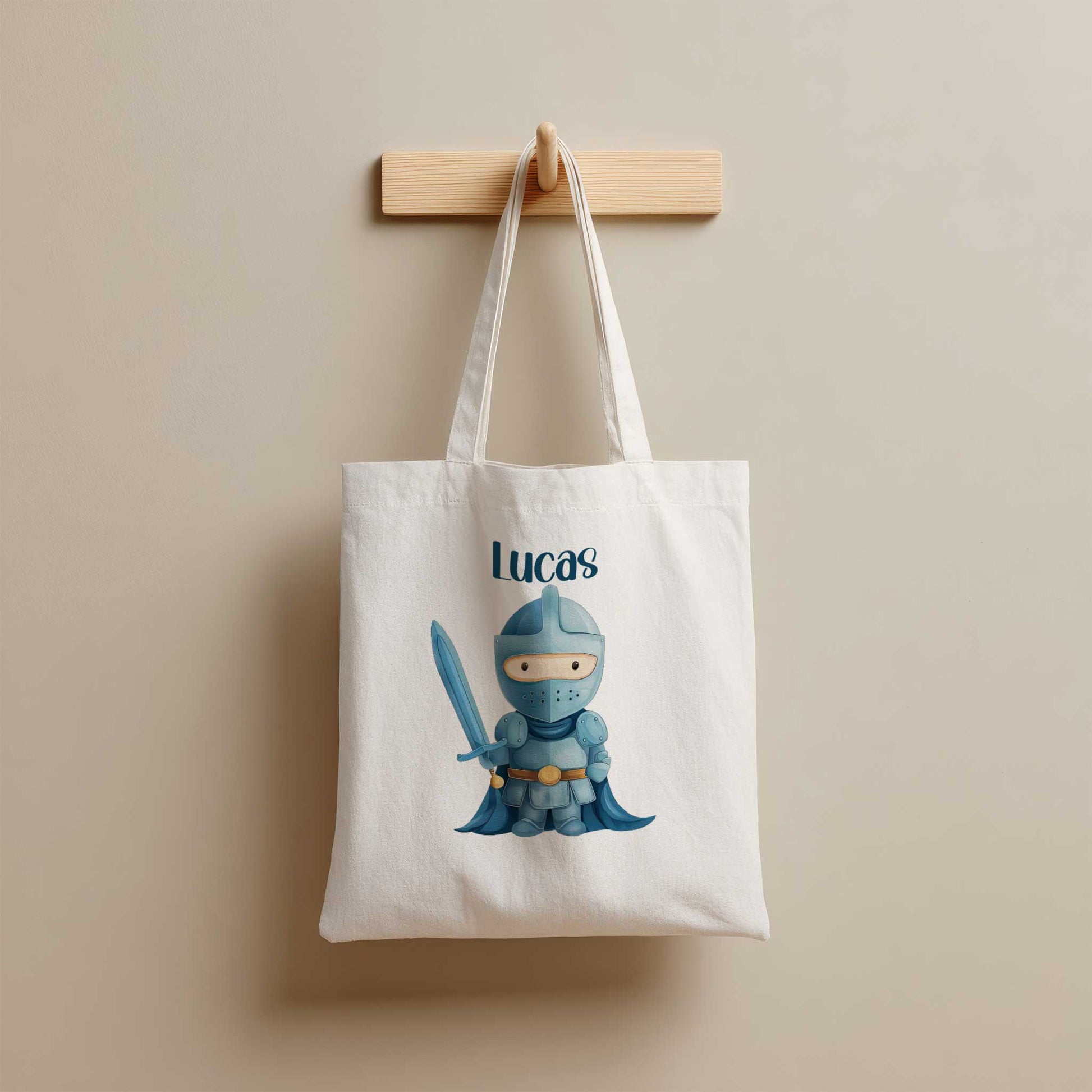 Witte totebag met de naam Lucas en een illustratie van een blauwe ridder met een zwaard, hangend aan een houten kapstok.
