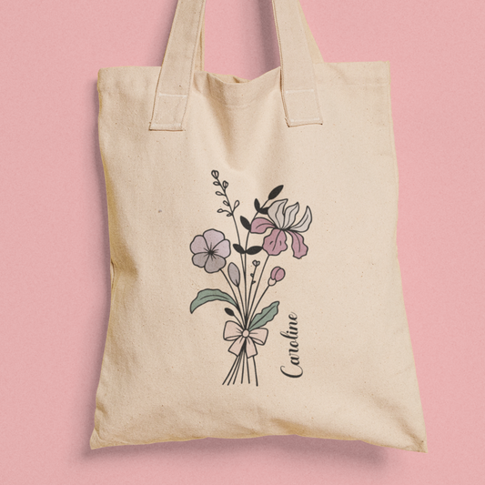 Totebag van katoen met bloemen lijntekening en naam Caroline, gefotografeerd tegen roze achtergrond