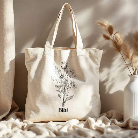 Katoenen totebag met zwarte bloemenlijntekening en naam Bibi, gefotografeerd op een bed met een lichtbeige sprei en kussens op de achtergrond.