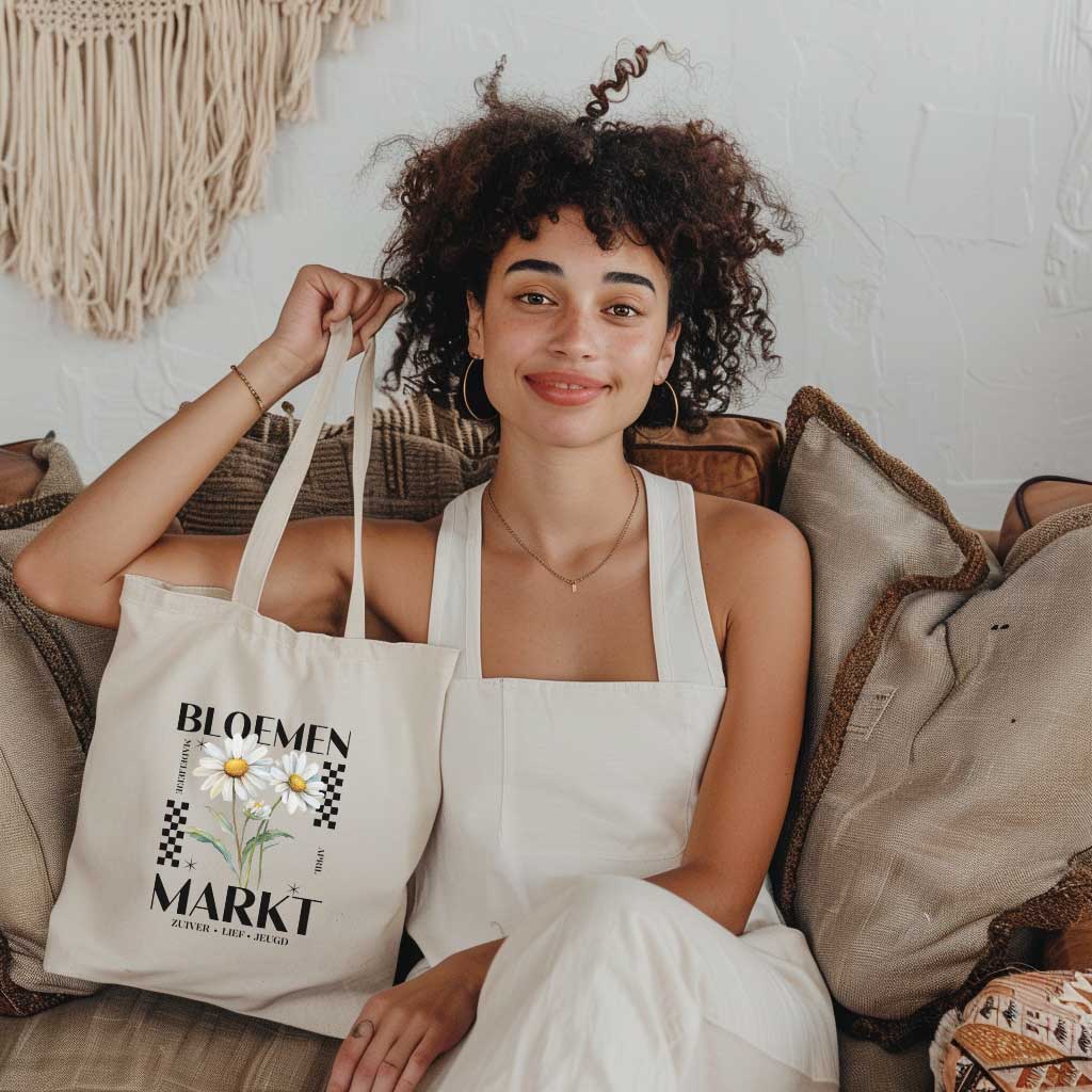 vrouw die op een bank zit en een totebag vasthoudt, totebag met  de tekst bloemenmarkt en een illustratie van een geboortebloem van de maand april, een madeliefje. Met onderaan de kernwoorden van deze maand : zuiver,lief,jeugd