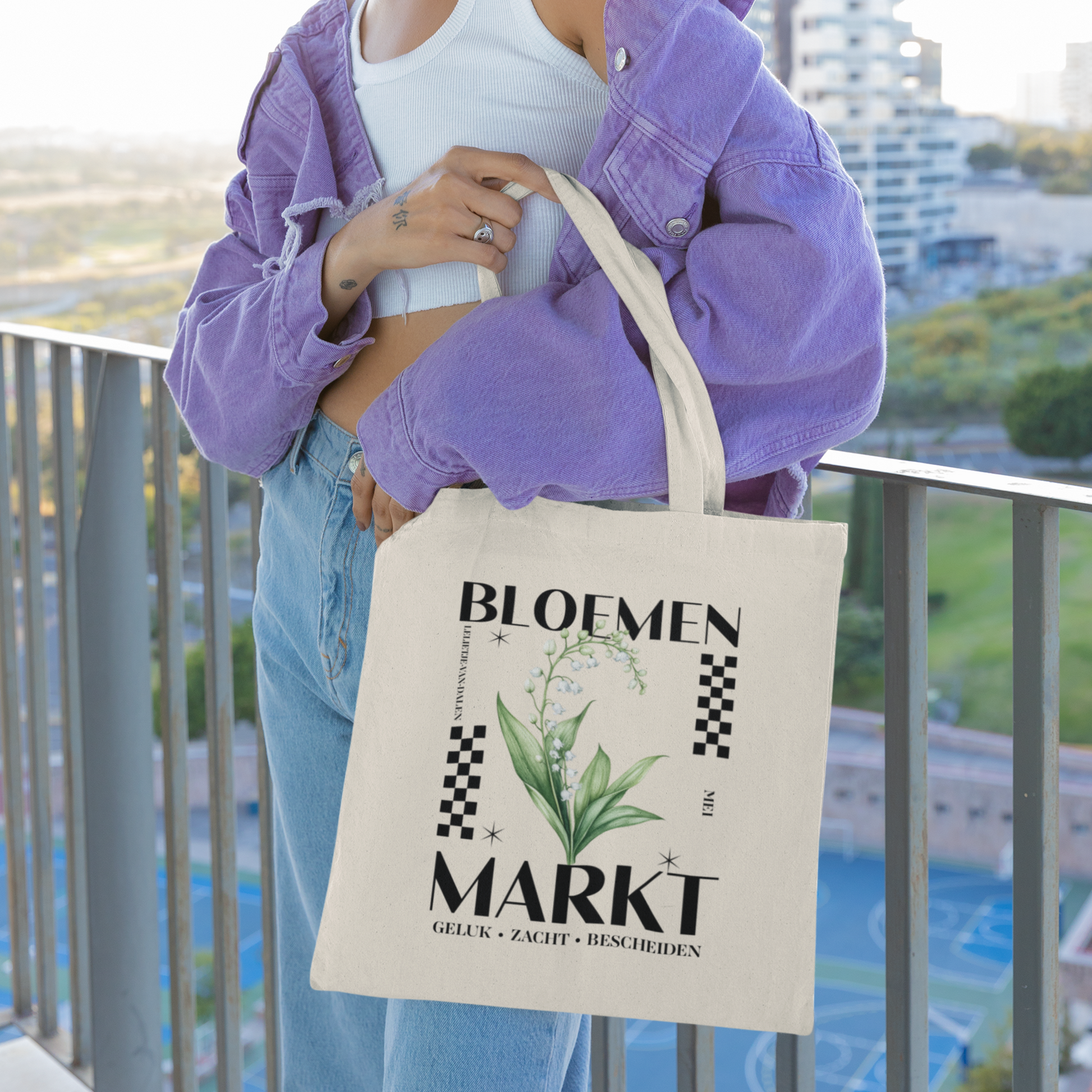 vrouw met paarse jas op een balkon met een totebag om haar hand, totebag met  de tekst bloemenmarkt en een illustratie van een geboortebloem van de maand mei, een Lelietje-van-dalen. Met onderaan de kernwoorden van deze maand : geluk,zacht,bescheiden.