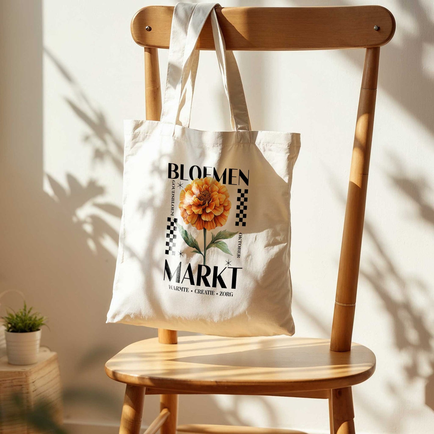totebag die aan een houten stoel hangt in het zonlicht. totebag met  de tekst bloemenmarkt en een illustratie van een geboortebloem van de maand oktober, een Goudsbloem. Met onderaan de kernwoorden van deze maand : warmte, creatie, zorg.