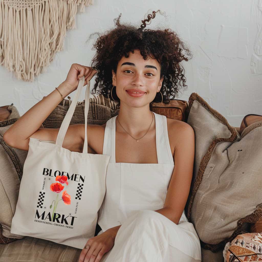 meisje die op een bank zit en een tote bag vasthoudt, totebag met  de tekst bloemenmarkt en een illustratie van een geboortebloem van de maand augustus klaprozen. Met onderaan de kernwoorden van deze maand : herinnering, troost, moed